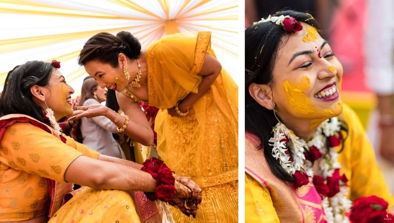 Two women in traditional Indian wedding attire exchanging garlands with smiles and laughter, with yellow and pink decorations in the background. One woman has yellow powder on her face and is wearing flowers around her neck.