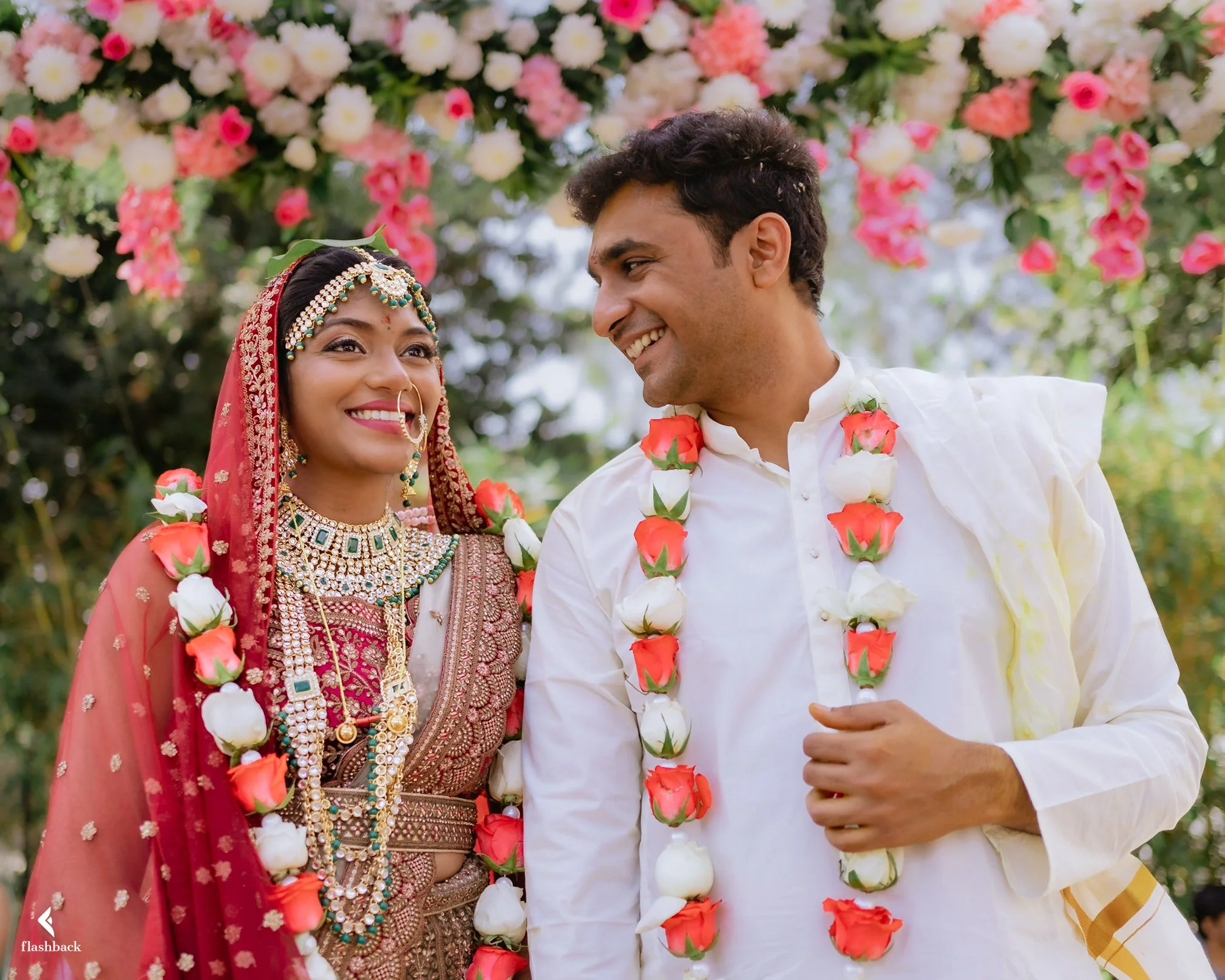 A newlywed Indian couple dressed in traditional wedding attire, adorned with flower garlands, smiling at each other, standing outdoors with a backdrop of pink and white flowers.