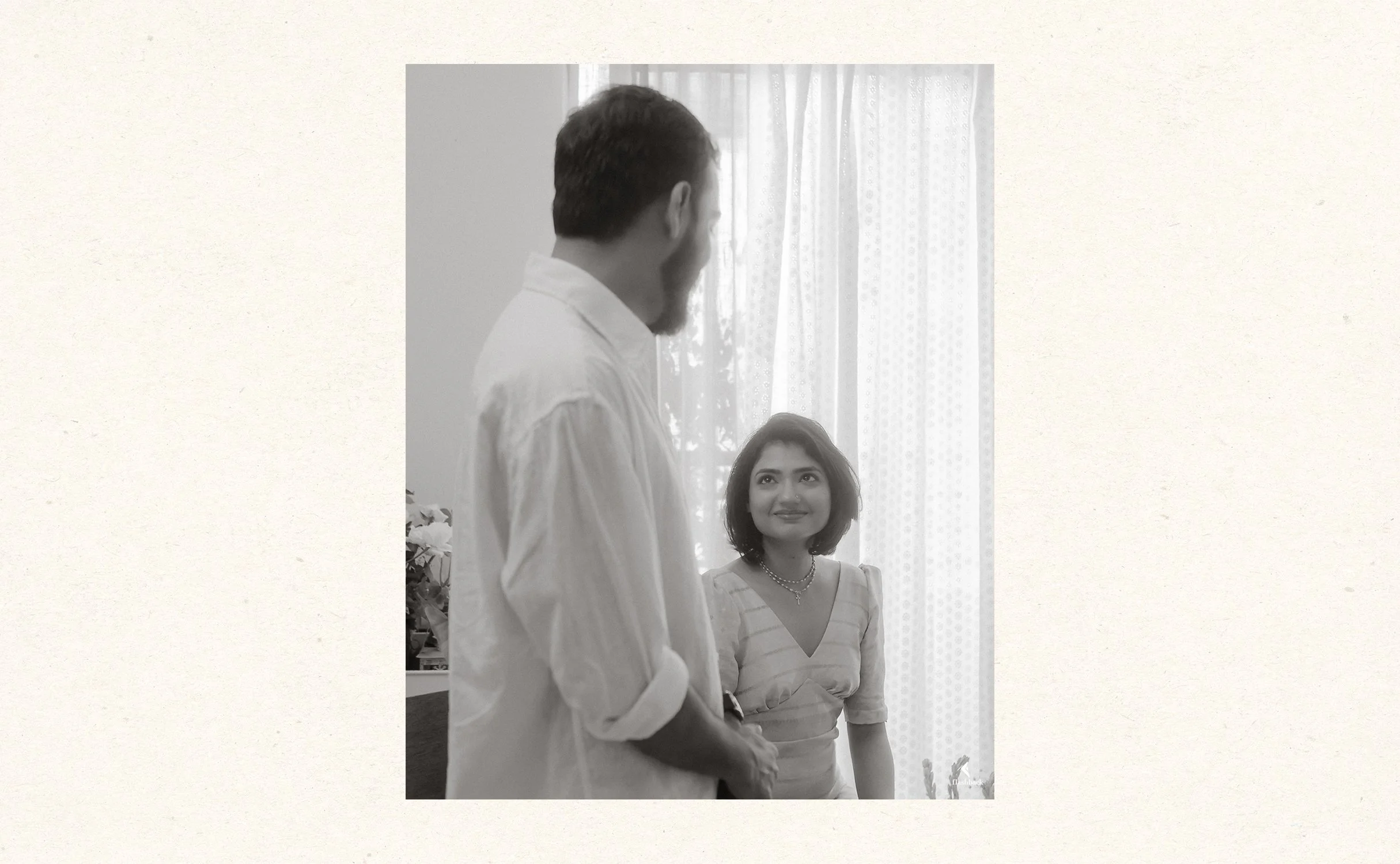 A man and woman indoors, with the man standing and facing the woman, who is looking up at him and smiling. The woman is wearing a striped top and necklace, while the man is in a light-colored shirt. There are curtains and plants in the background.