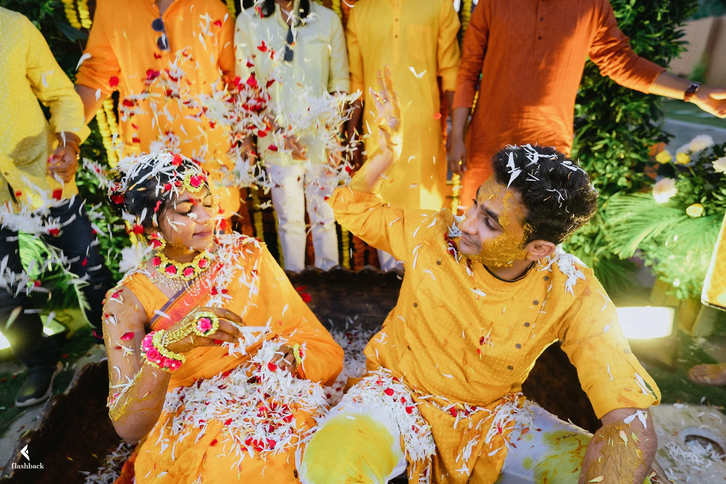 A couple celebrating a traditional Indian wedding, surrounded by friends, with flower petals in the air, wearing bright orange and yellow attire.