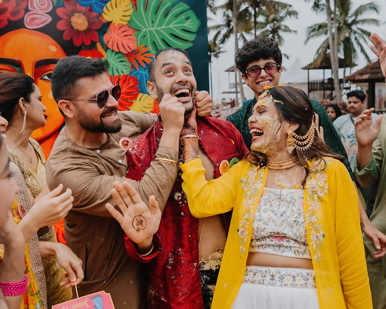 A group of people celebrating and smiling at an outdoor event, with a man and woman covered in yellow and white powder, wearing traditional clothing and jewelry, amidst a festive atmosphere with colorful decorations and palm trees in the background.