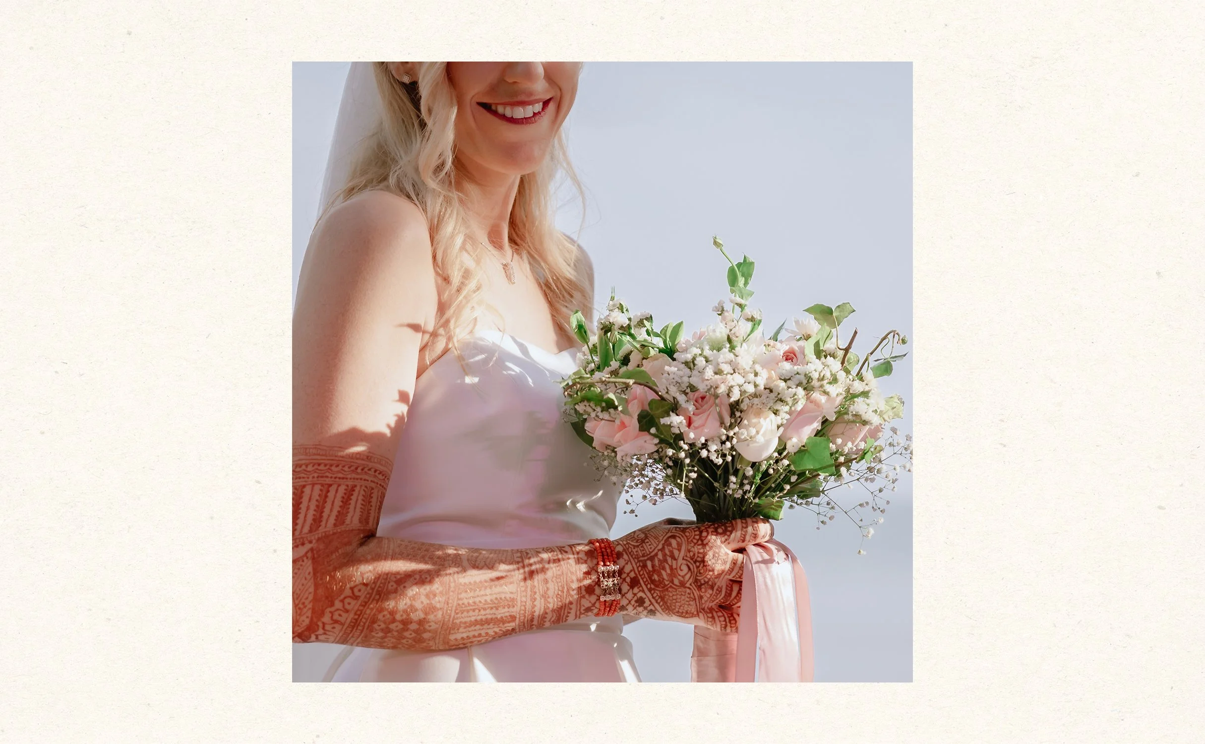 A smiling bride with long blonde hair holding a bouquet of pink and white flowers, wearing a white sleeveless wedding dress and intricate henna designs on her arms, standing outdoors against a clear sky.