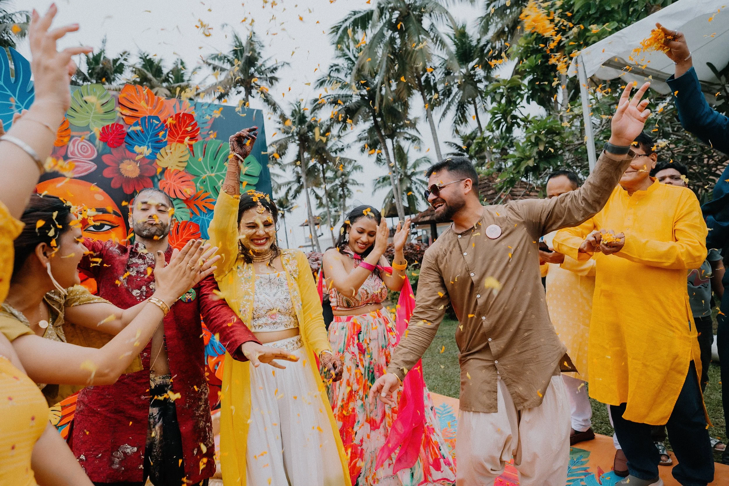 Group of people celebrating at an outdoor event with colorful decorations and yellow flower petals falling from above, surrounded by palm trees.