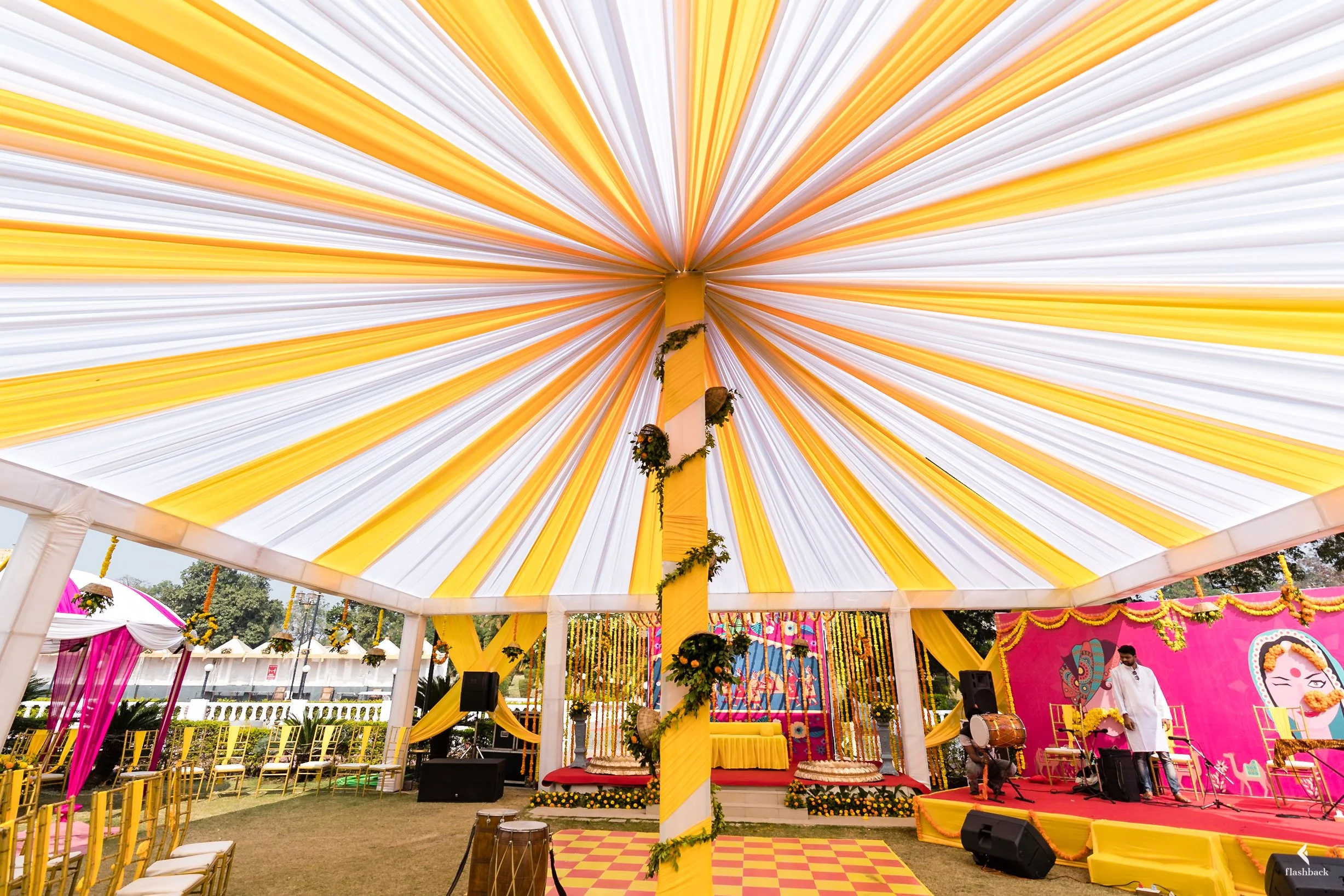 Decorated event tent with yellow and white canopy, floral and music setup, pink backdrop, and seating arrangements.