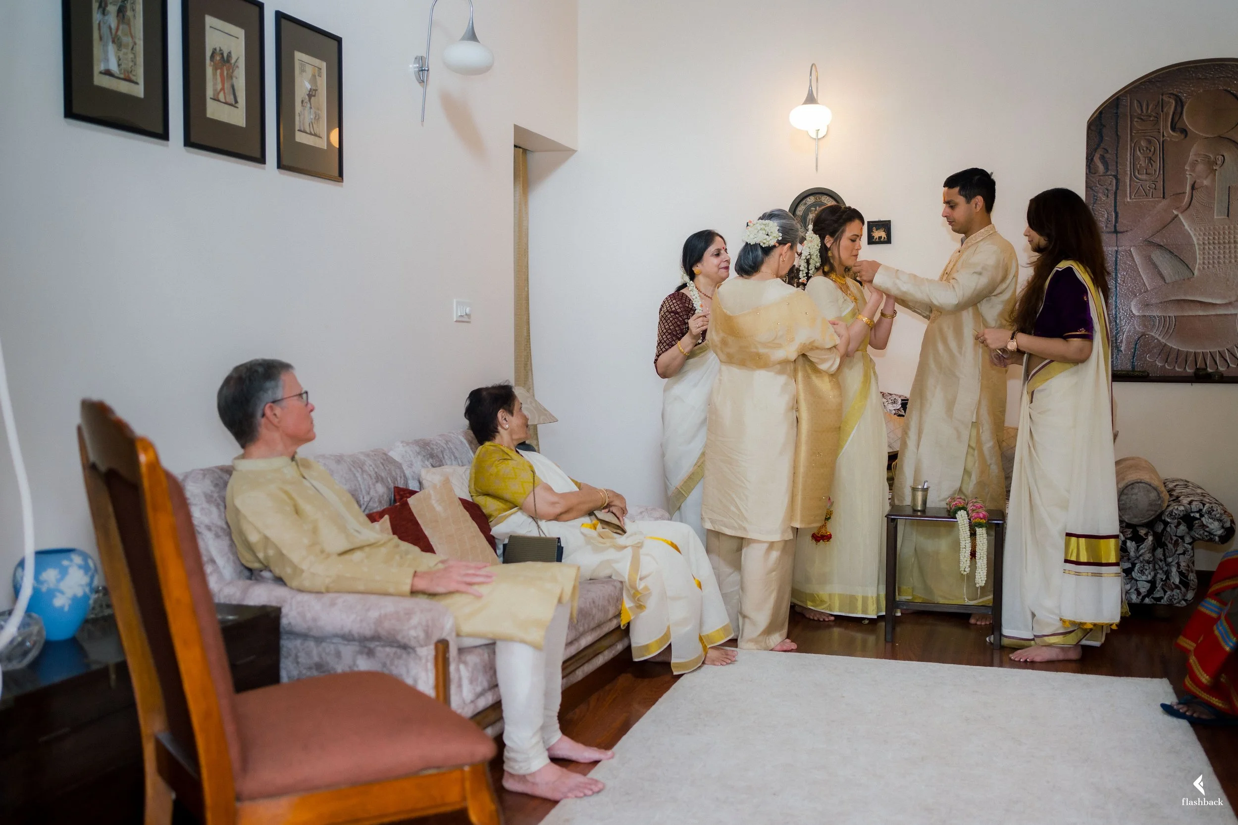 A traditional Indian family gathering in a living room. Several women and men dressed in traditional attire are participating in a ceremonial ritual while three elderly women sitting on a sofa observe. The room is decorated with framed artwork and a