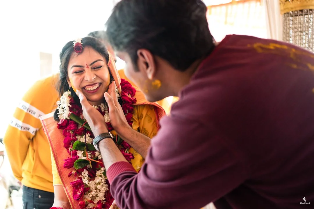 A woman dressed in traditional Indian attire with flowers in her hair is smiling joyfully as a man applies turmeric to her face during a celebration.