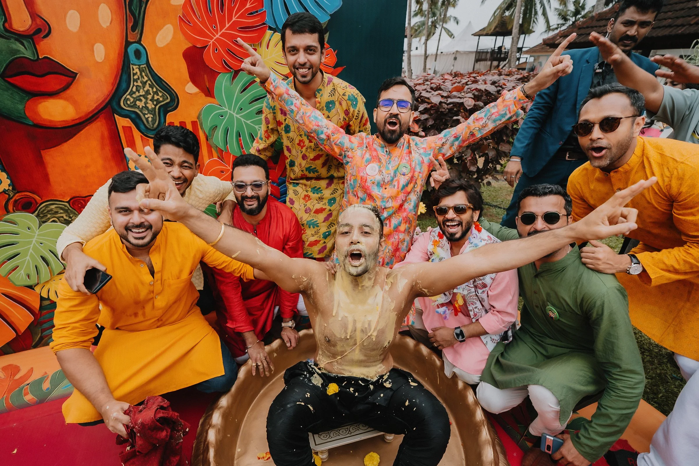 A group of men celebrating Holi, the Indian festival, in front of a colorful backdrop with abstract face and leaf designs. One man is covered in splashes of color, sitting in a traditional Indian clay pot, with others around him joyfully participatin