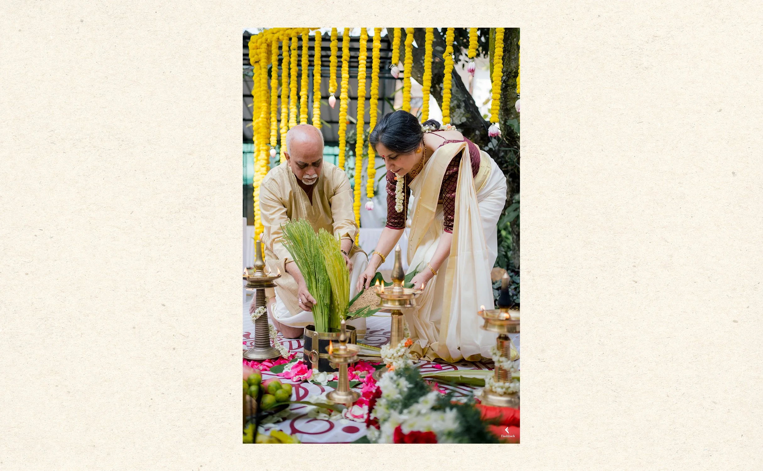 Two people, a man and a woman, are participating in a traditional Indian religious or cultural ceremony outdoors. They are kneeling and handling plants or offerings on a decorated surface with items like lamps, flowers, and fruits around them. Yellow