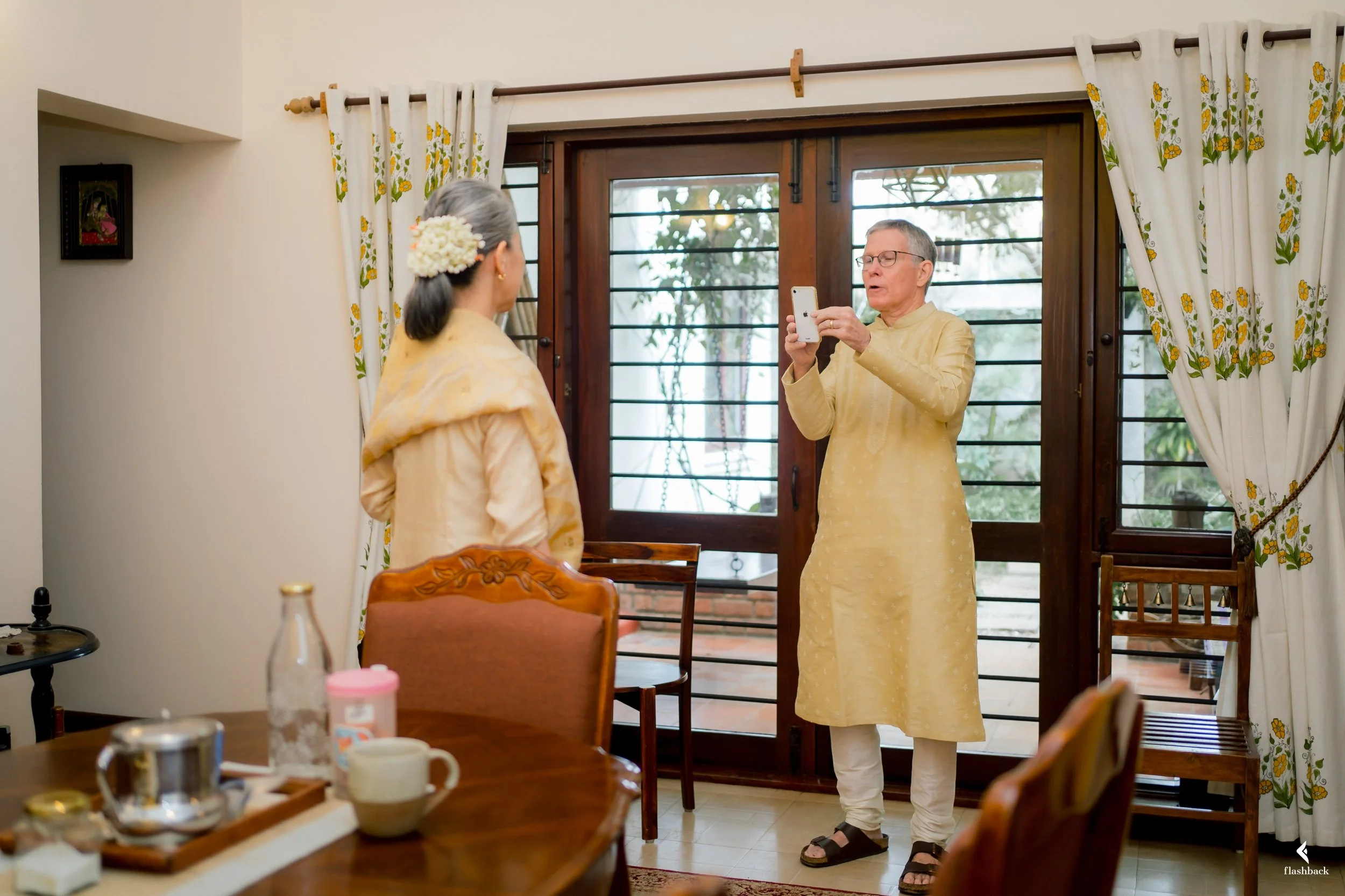 An elderly man in traditional Indian attire is taking a photo of a woman in similar attire inside a home with wooden furniture and floral curtains.