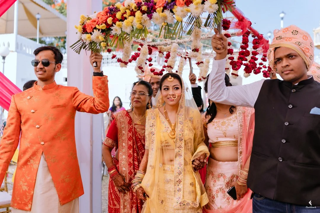 Indian bride in yellow traditional attire with jewelry and nose ring, surrounded by family members at a wedding, under a floral canopy.