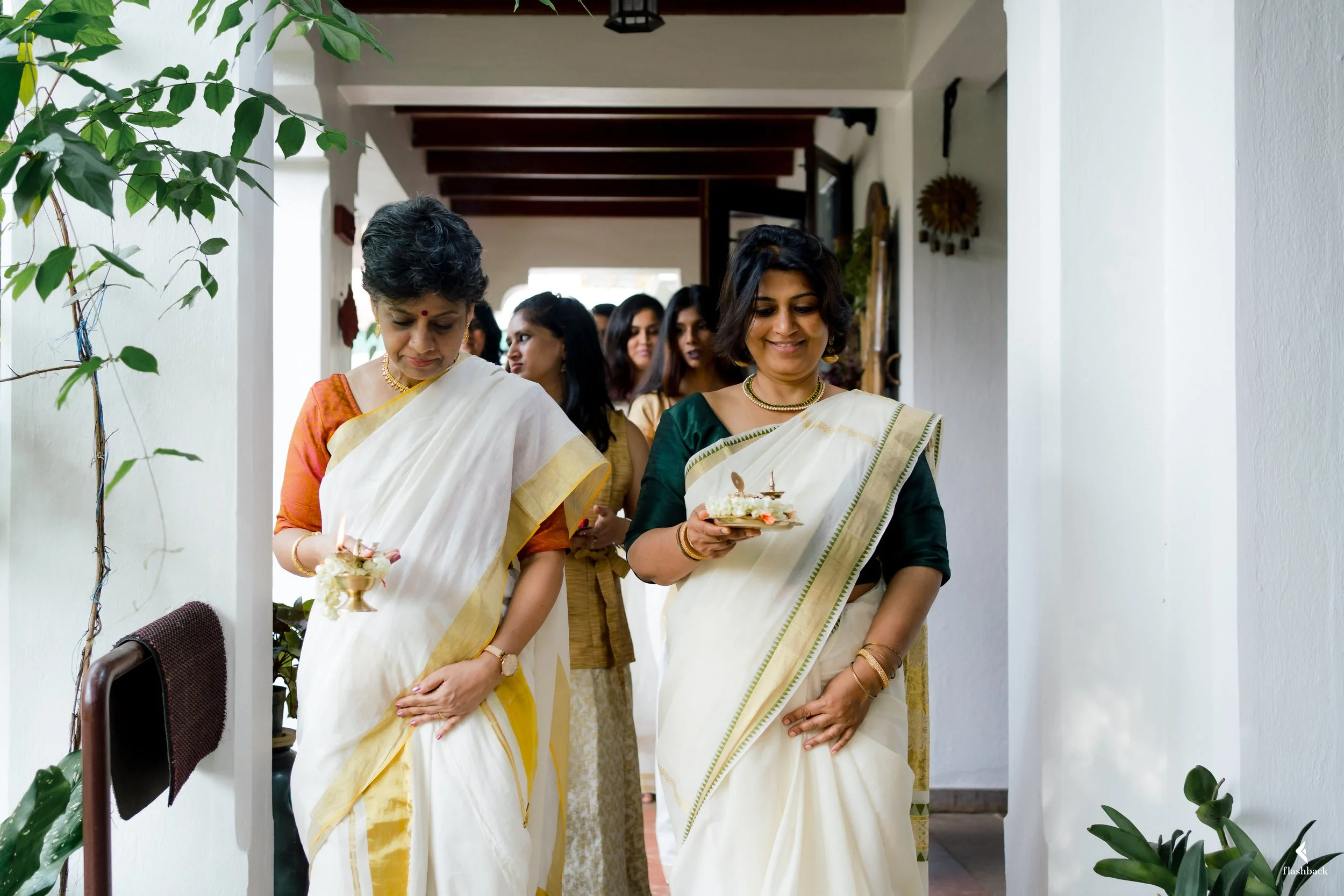 Two women in traditional Indian sarees, carrying small decorated plates with lamps and flowers, participate in a religious or cultural ceremony inside a house, with other women in the background.