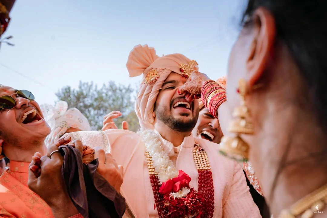 Indian groom in traditional wedding attire, laughing and being blessed by family members during an outdoor wedding celebration.