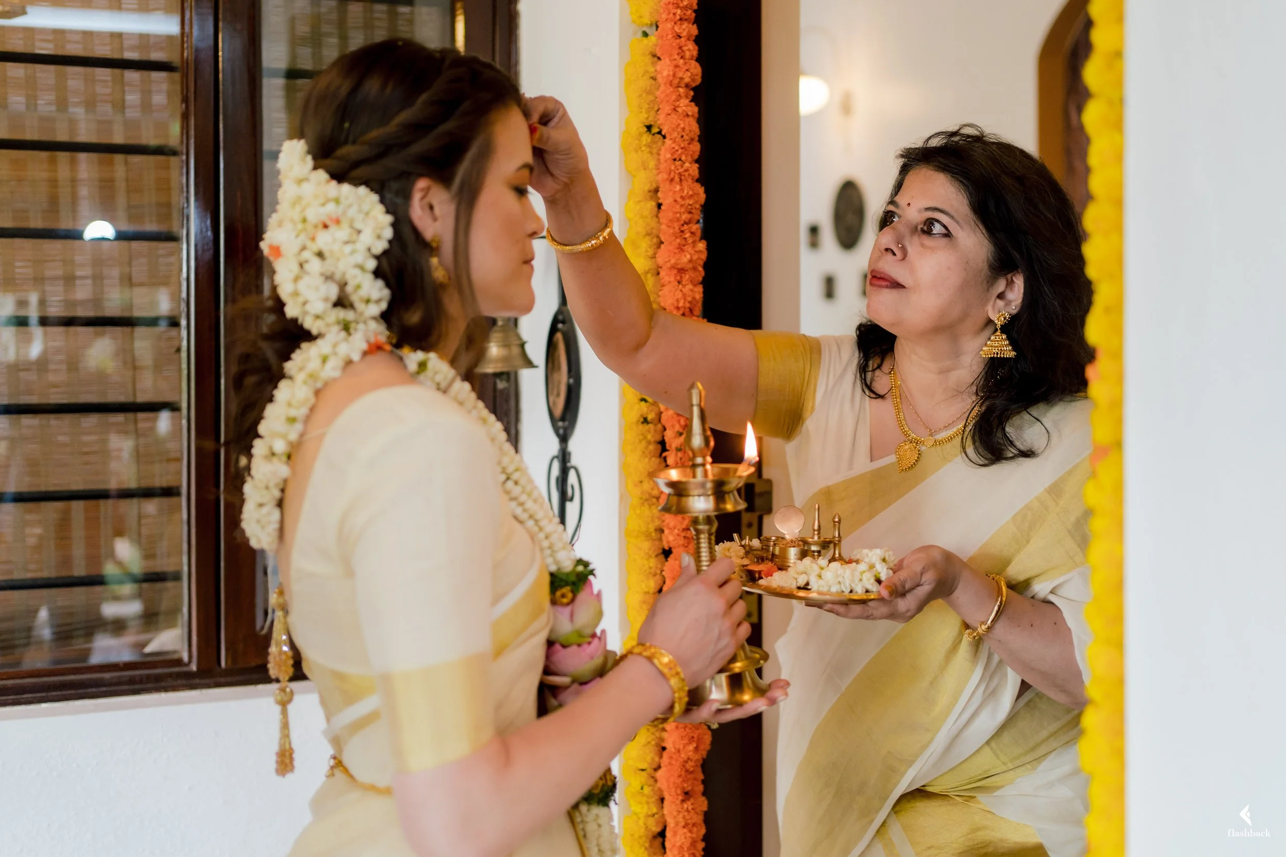 A woman dressed in traditional Indian attire with flowers in her hair participates in a religious ceremony while another woman performs a ritual, holding a lamp and tray with offerings, near a decorated doorway.