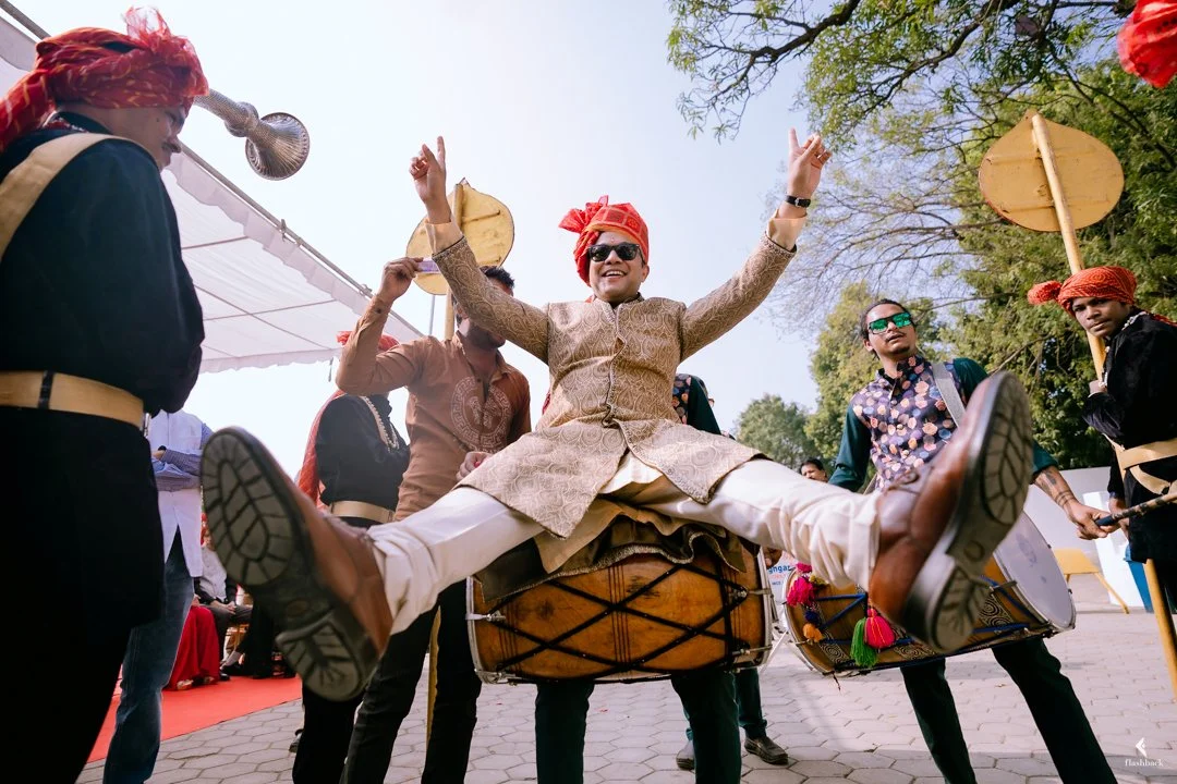 A young man wearing traditional Indian attire, sunglasses, and a headscarf, jumping with a joyful expression on a large drum during a cultural celebration, surrounded by people dressed in colorful traditional clothing and playing drums beneath a clea