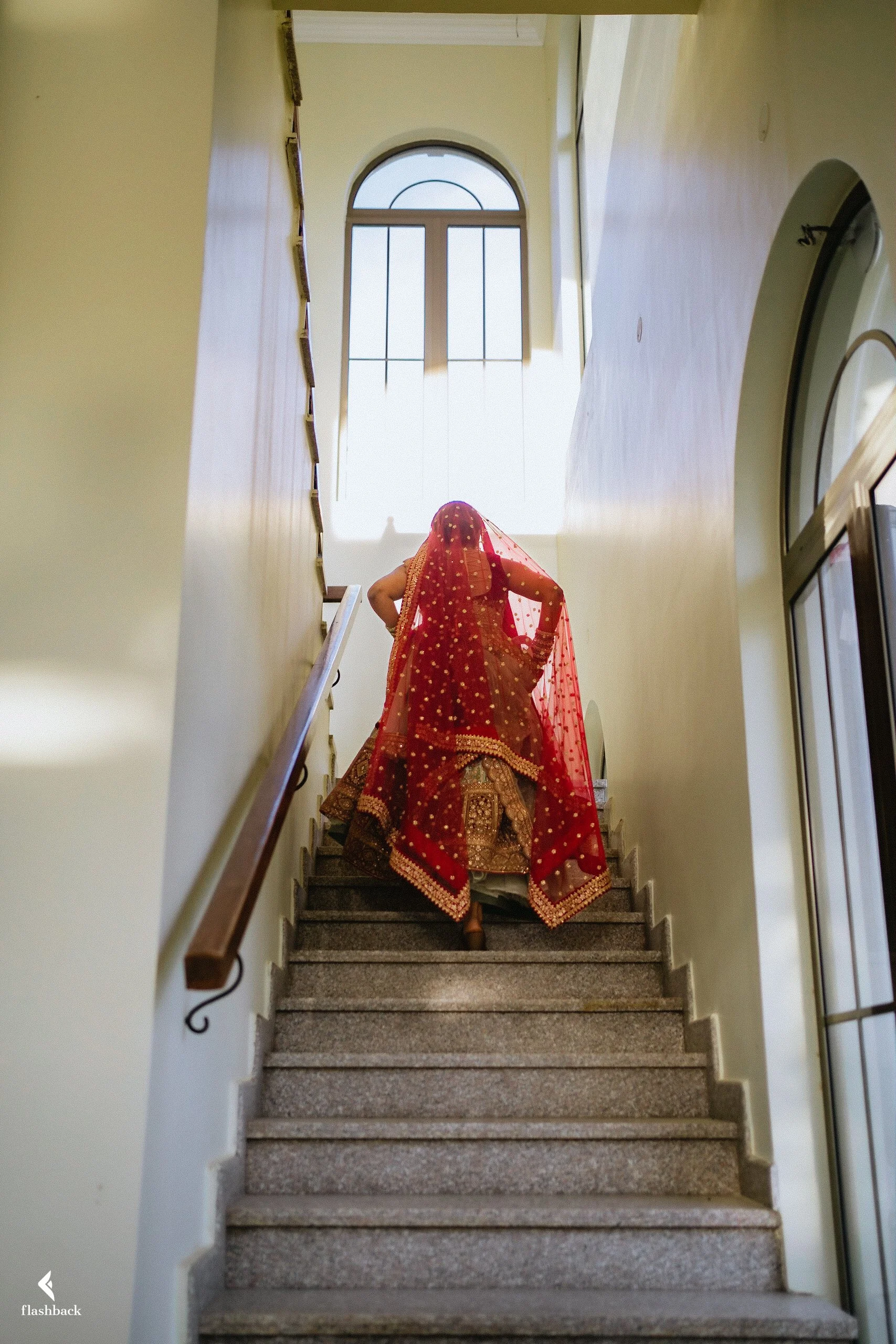 A woman dressed in traditional Indian bridal attire, including a red saree with gold embroidery and a red veil, walks up a staircase near a large arched window.