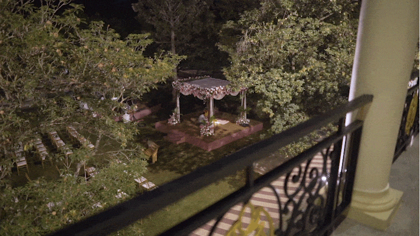 Nighttime view of a decorated outdoor wedding or event area as seen from a balcony, with a floral arch, chairs, and trees.