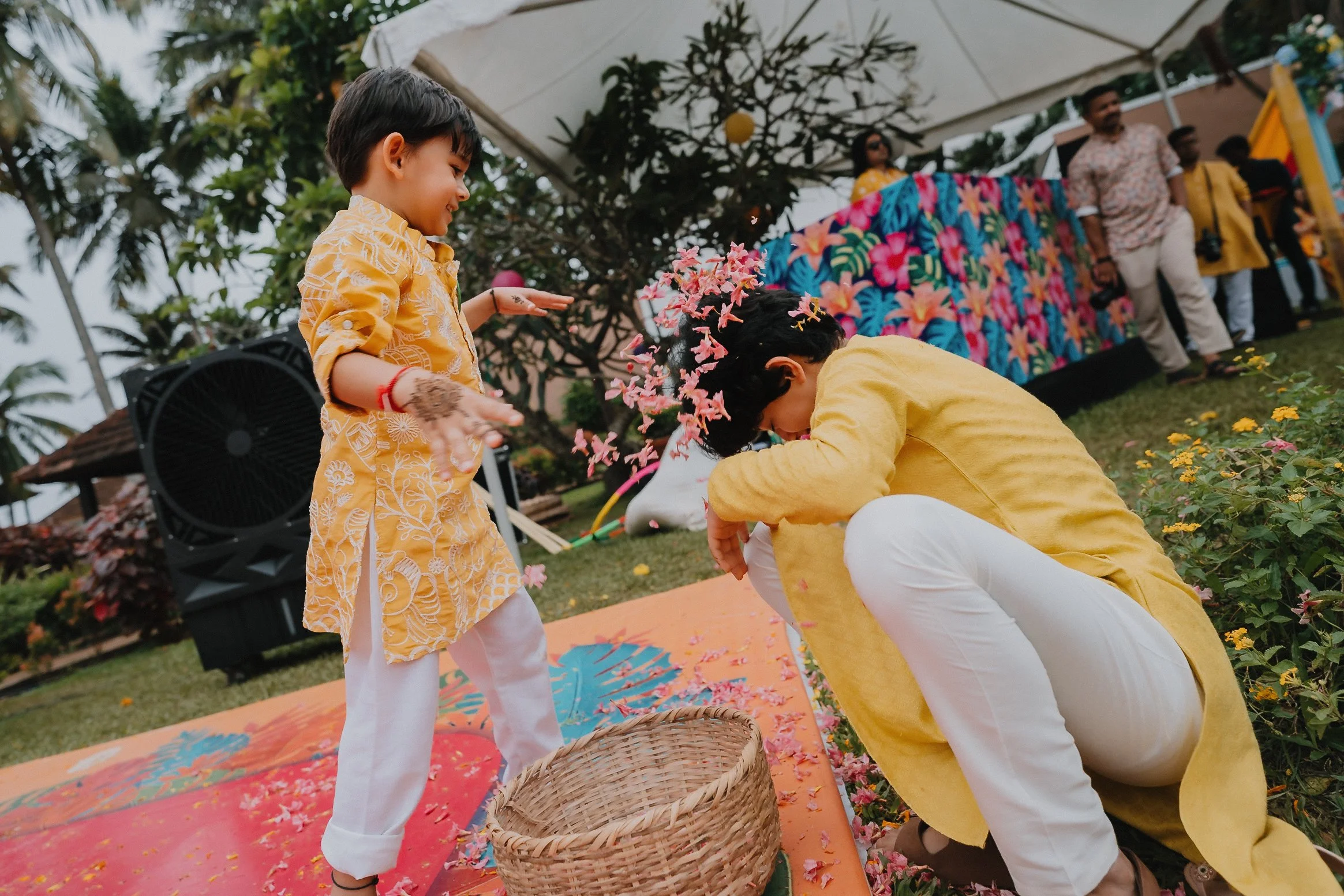 Children celebrating at an outdoor event with flower petals, wearing yellow and white traditional clothing, under a canopy with decorated backdrop.