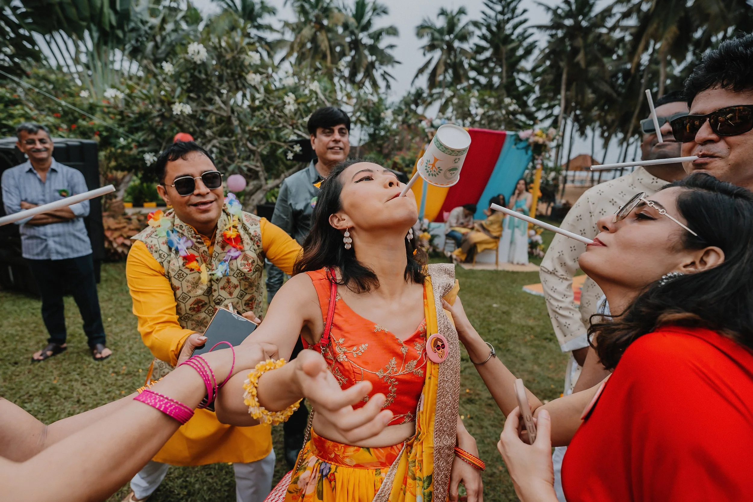 Group of people celebrating at an outdoor festival, blowing party blowers and wearing colorful outfits, with palm trees and decorations in the background.