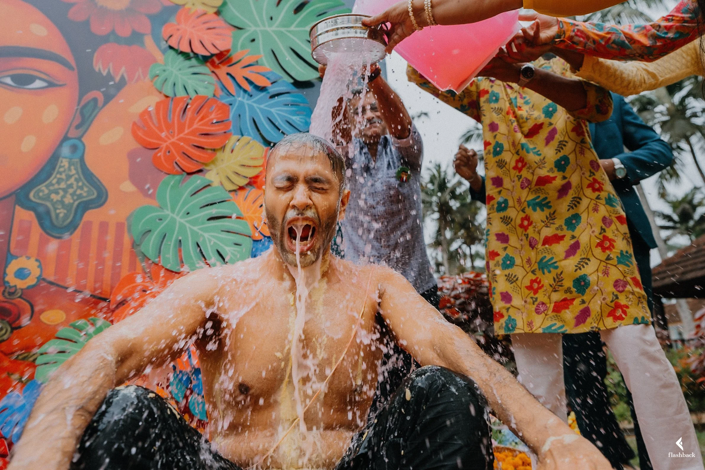 People participating in a traditional ritual involving pouring water over a person's head, with a colorful decorative background featuring leaves and artwork.