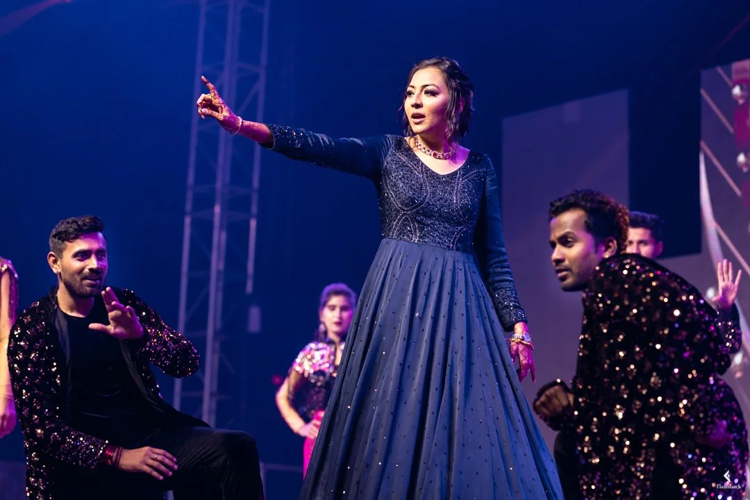 A woman in a long, dark blue, sparkly dress performs on stage, accompanied by backup dancers.