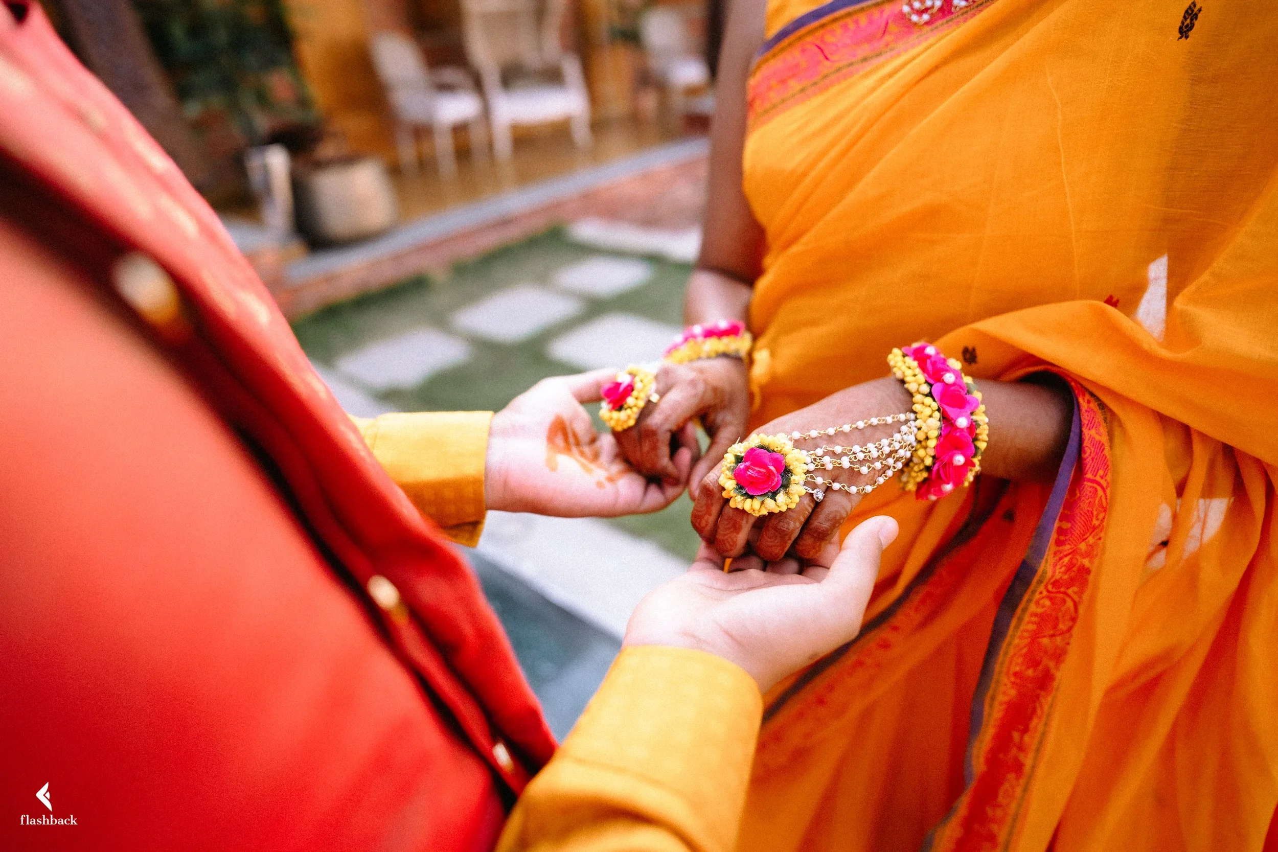 A close-up of a traditional Indian wedding ceremony showing a couple holding hands, with the bride wearing yellow and pink floral jewelry on her hands and wrists, and a yellow saree with pink accents. The groom is dressed in orange and gold attire.