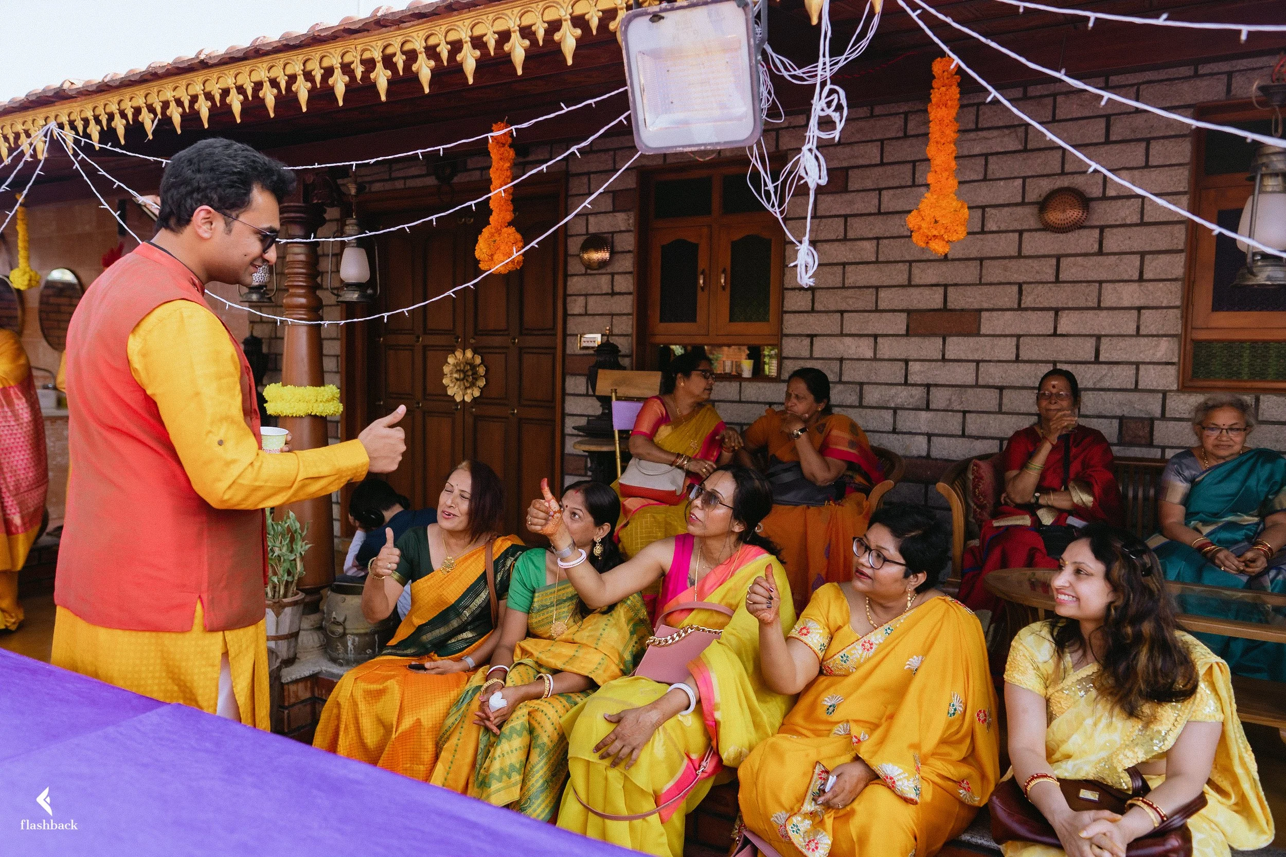 A group of women wearing traditional Indian sarees sitting and chatting at a festive gathering at home. A man in a yellow and orange kurta stands in front of them, smiling and engaging with the group. Festive decorations such as marigold flower garla