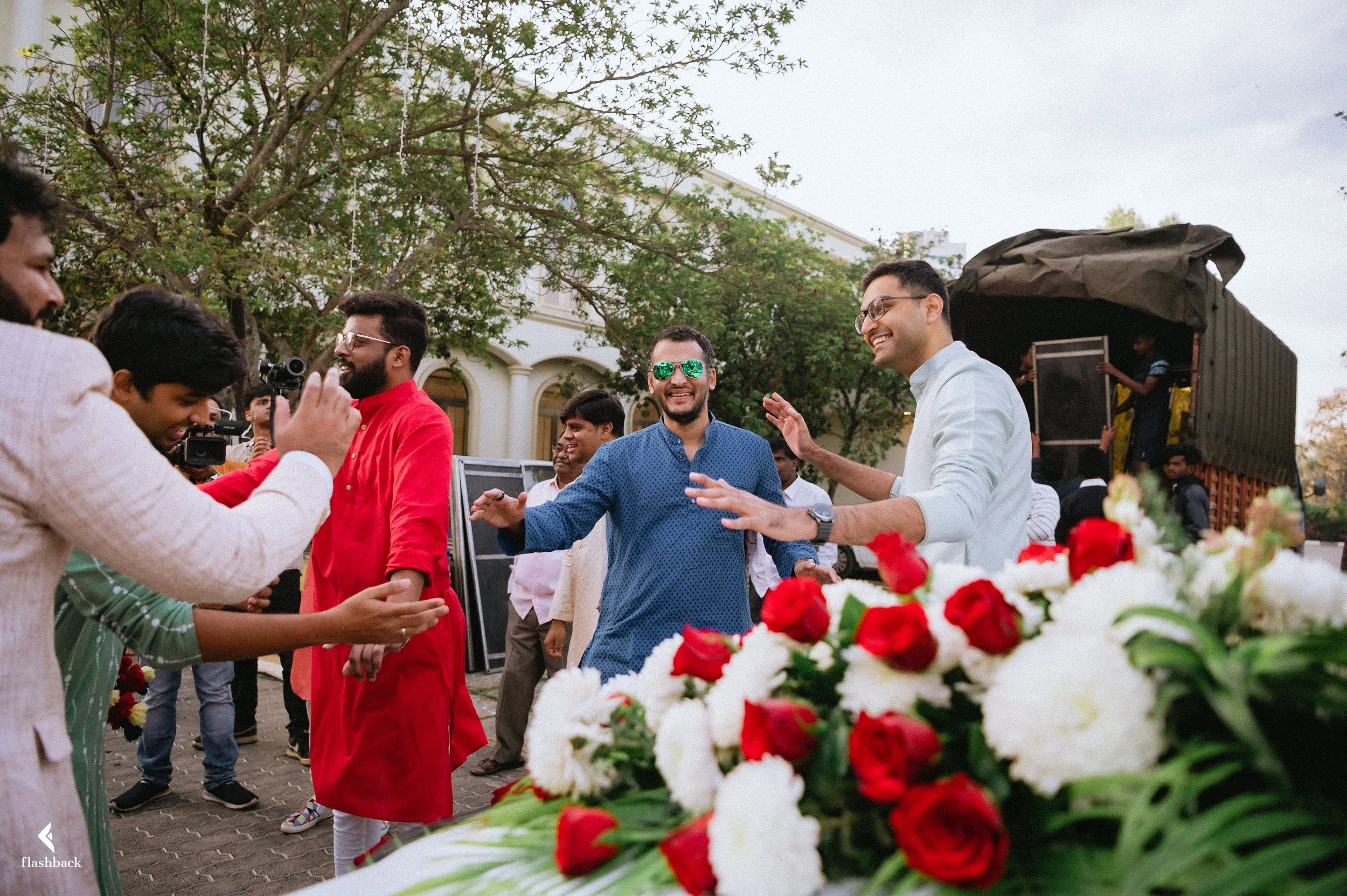 A group of men smiling and dancing at an outdoor celebration, with a flower arrangement in the foreground and a truck in the background.