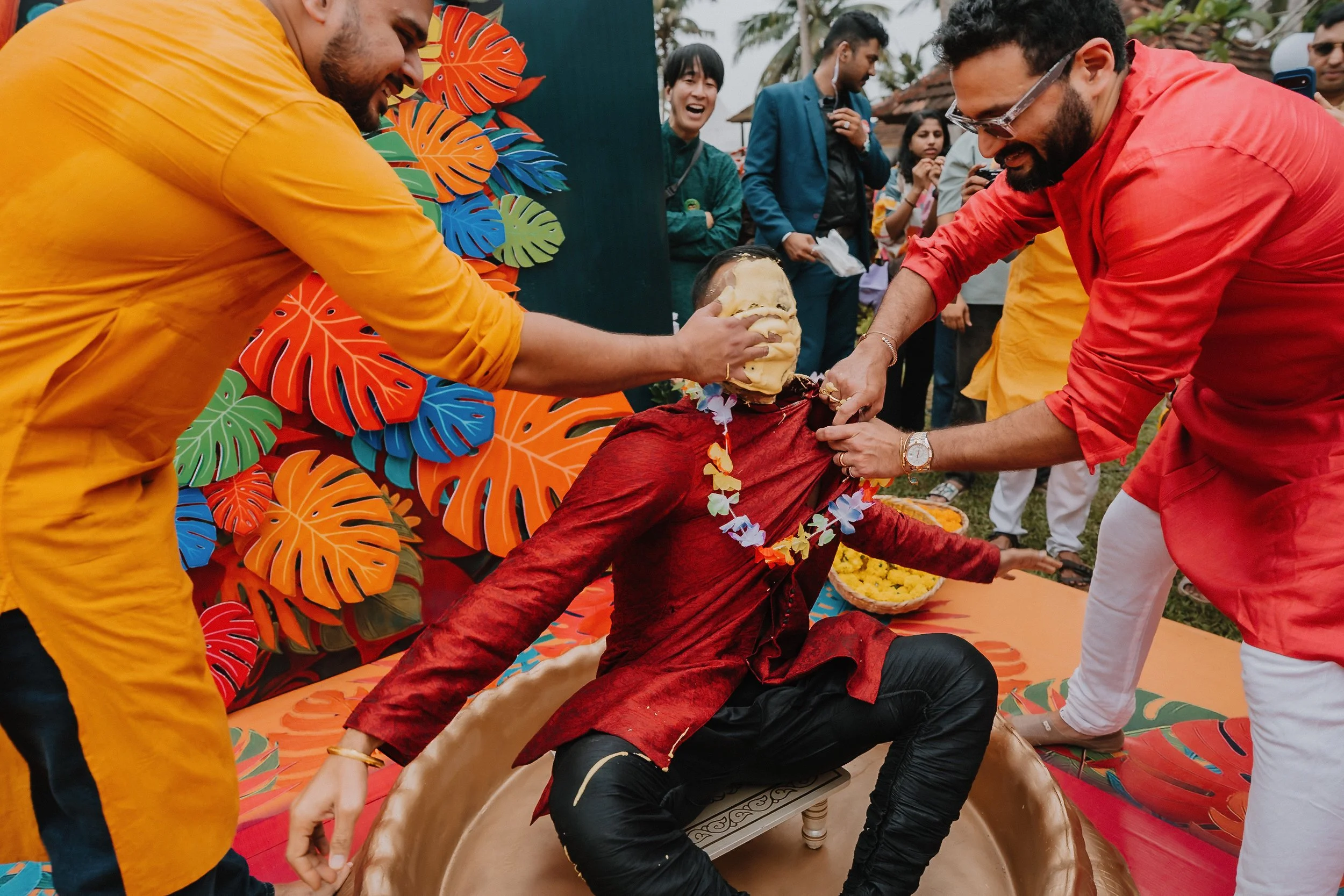 A group of people celebrating a Hindu ritual where a man is taking a sacred bath with a coconut, amid vibrant tropical decorations and onlookers smiling.