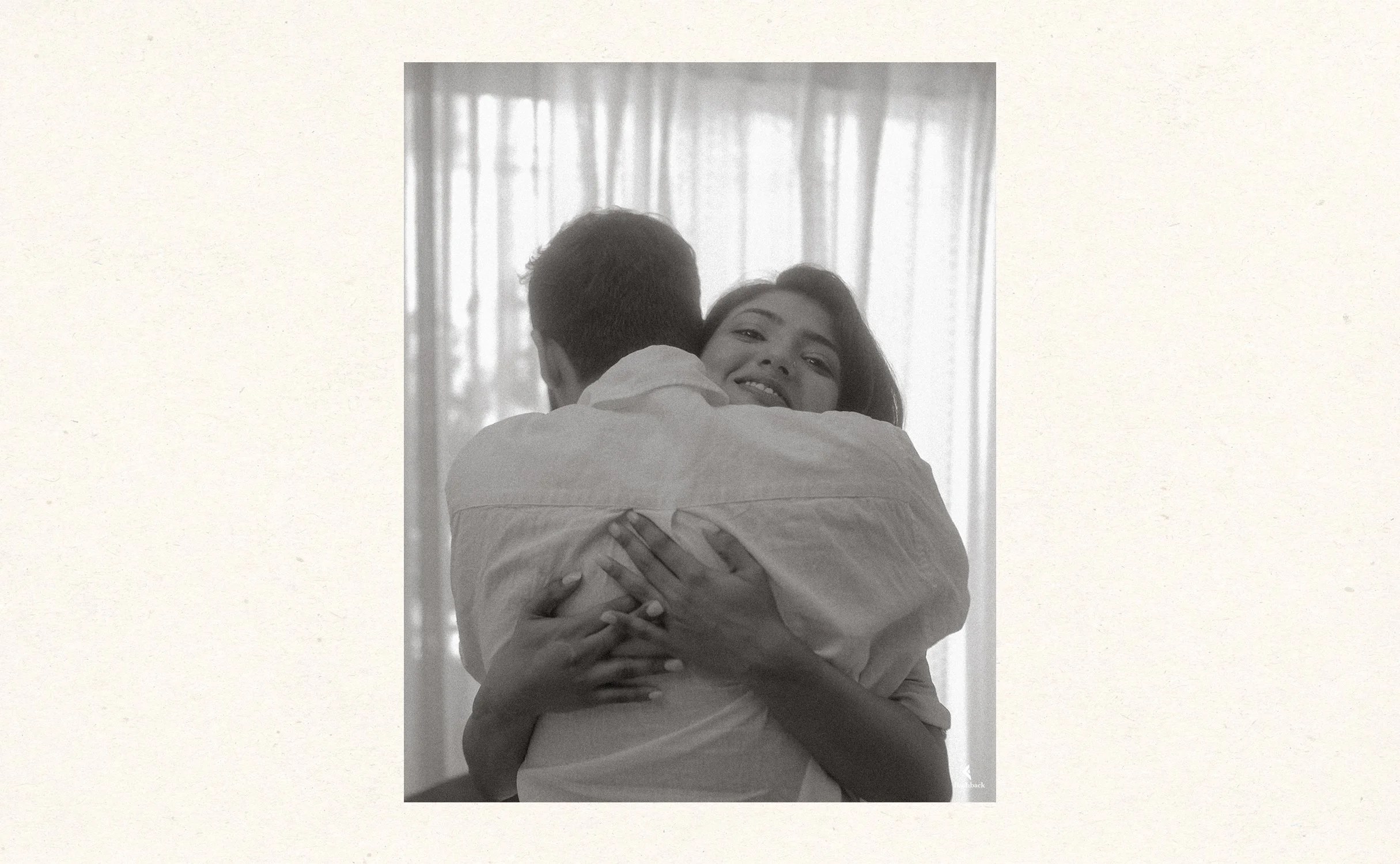 A black and white photo of a woman embracing a man, smiling and looking at the camera, with a background of curtains.
