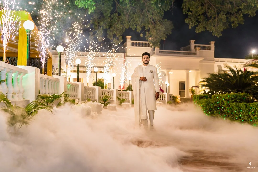 Man dressed in traditional Indian attire standing outdoors at night with fireworks, sparklers, and decorative lighting, surrounded by lush plants and a mist or fog effect.