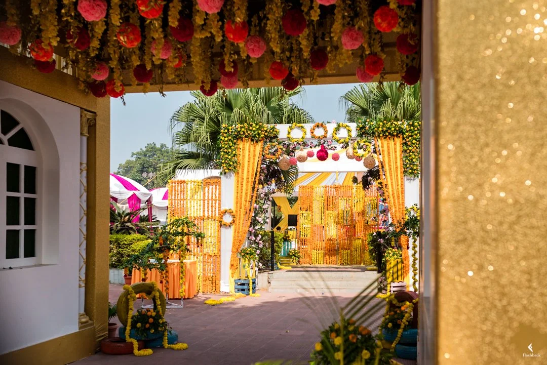 Colorful wedding decorate with yellow, pink, and orange flowers, with an arch and drapes, set outdoors under palm trees.