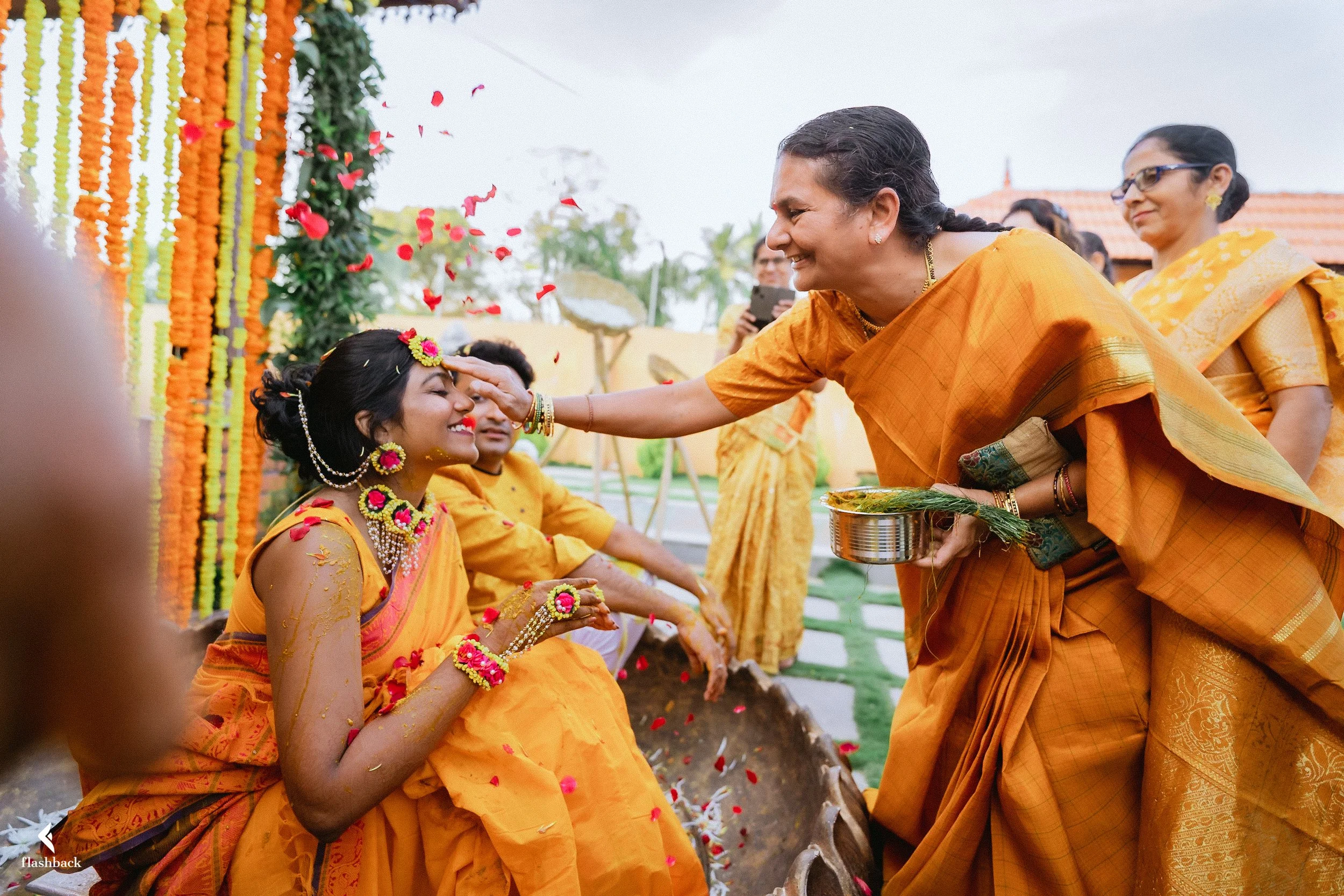 An Indian woman in orange saree is blessing a young woman in similar attire at a traditional ceremony, with others in yellow sarees observing nearby.