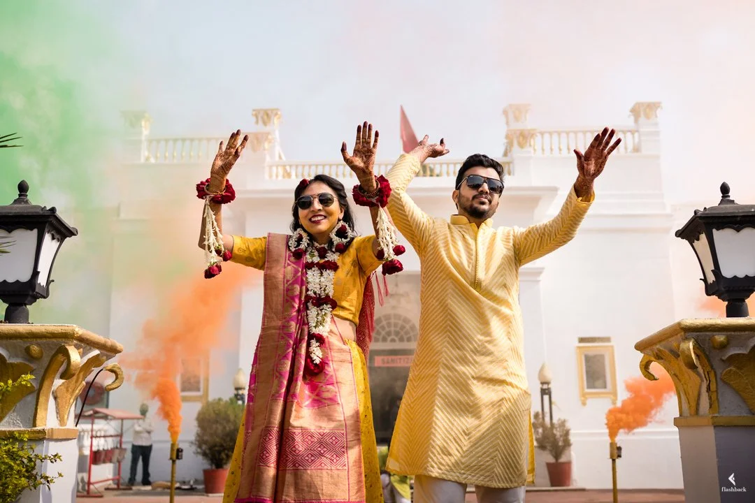 A smiling couple in traditional Indian attire celebrating outdoors at a wedding or festival, with smoke and decorative elements in the background.