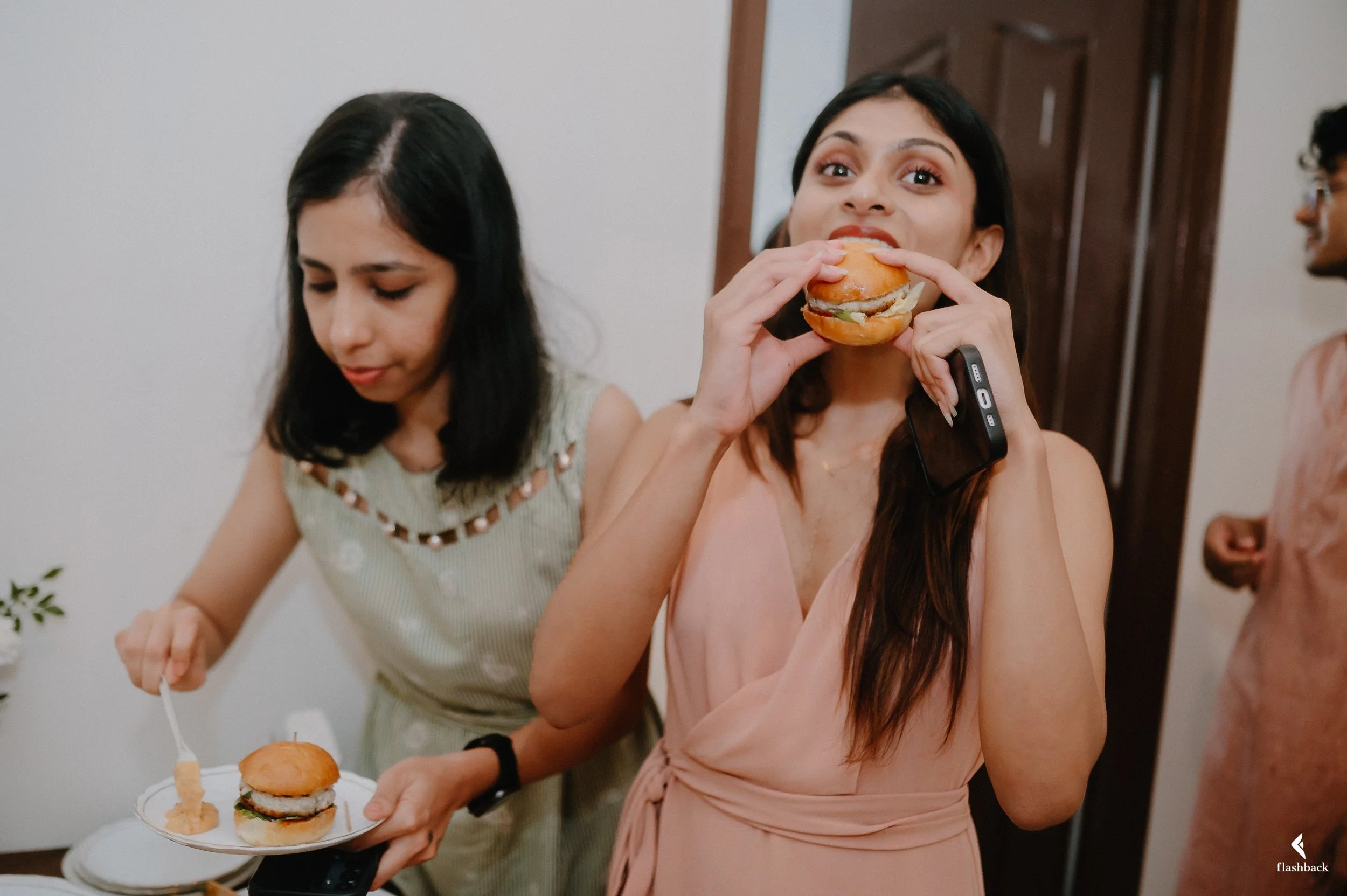 A woman in a peach dress is eating a hamburger at a social gathering, holding a mobile phone in her other hand. Next to her, another woman in a light-colored dress is serving herself a hamburger. There are other people in the background.