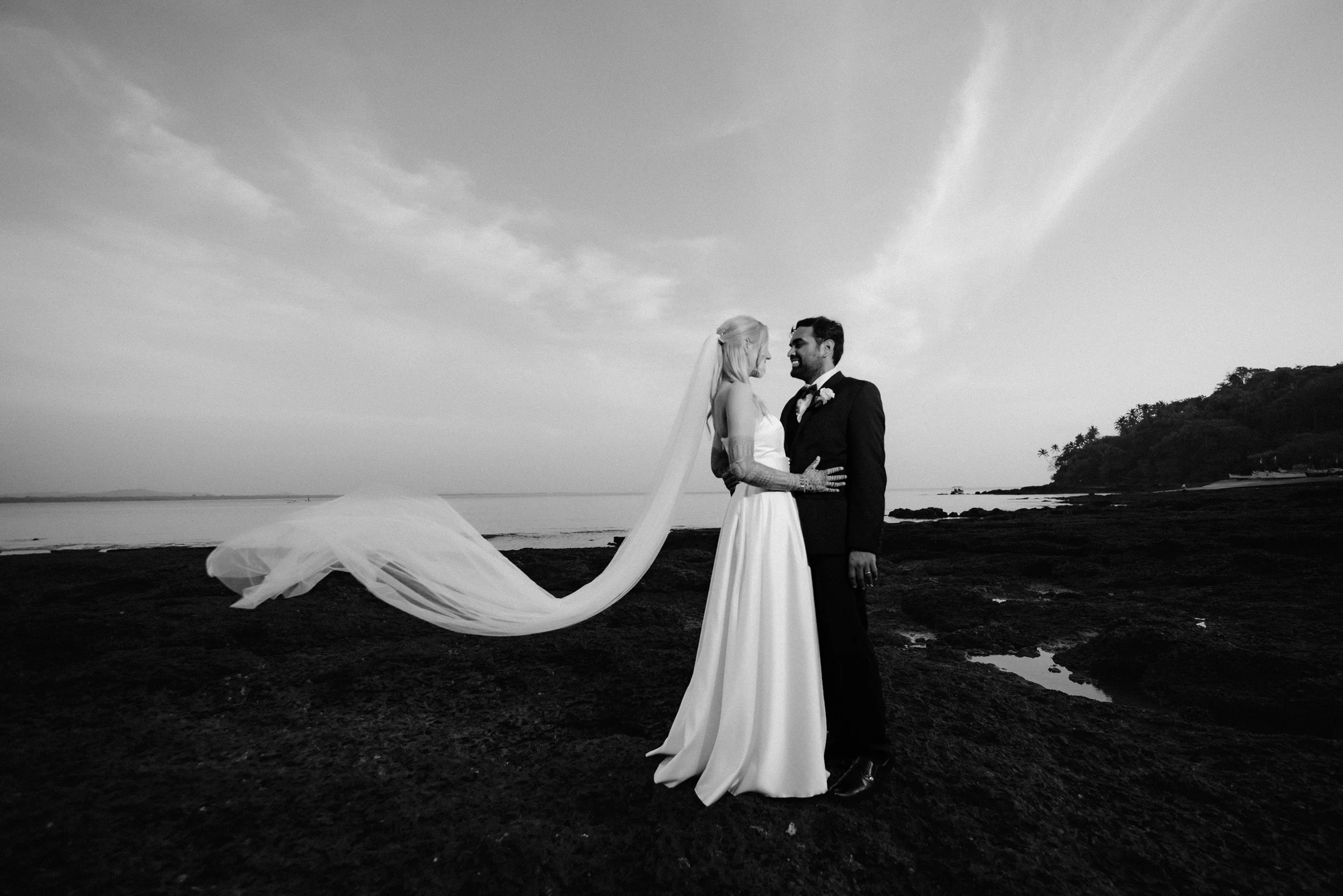 Black and white photo of a bride and groom on a beach, facing each other, holding hands, with water and a distant shore in the background, and the bride's veil flowing behind her.