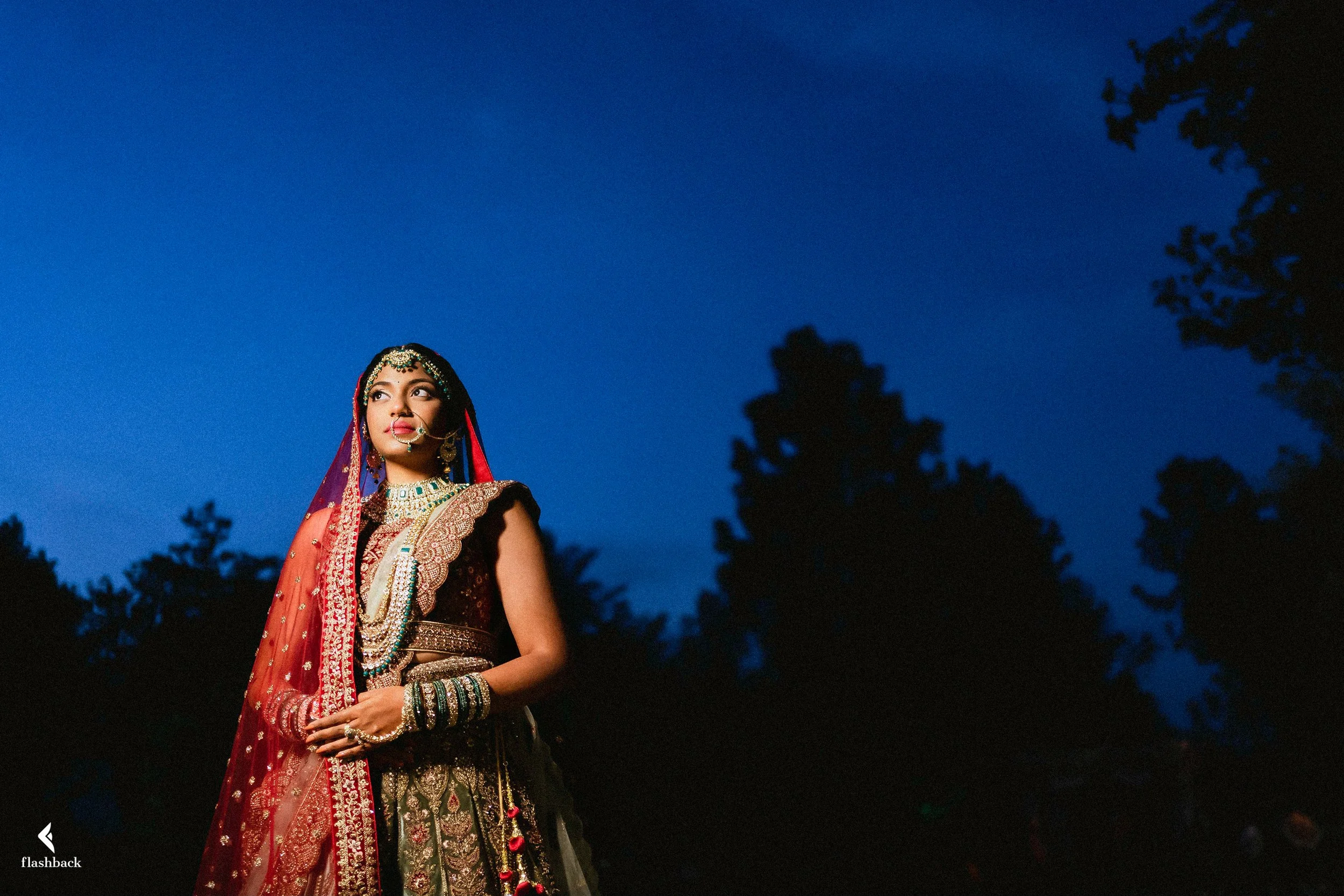 A woman dressed in traditional Indian bridal attire standing outdoors at dusk, adorned with jewelry and a red and gold embroidered outfit, with a deep blue sky and silhouettes of trees in the background.