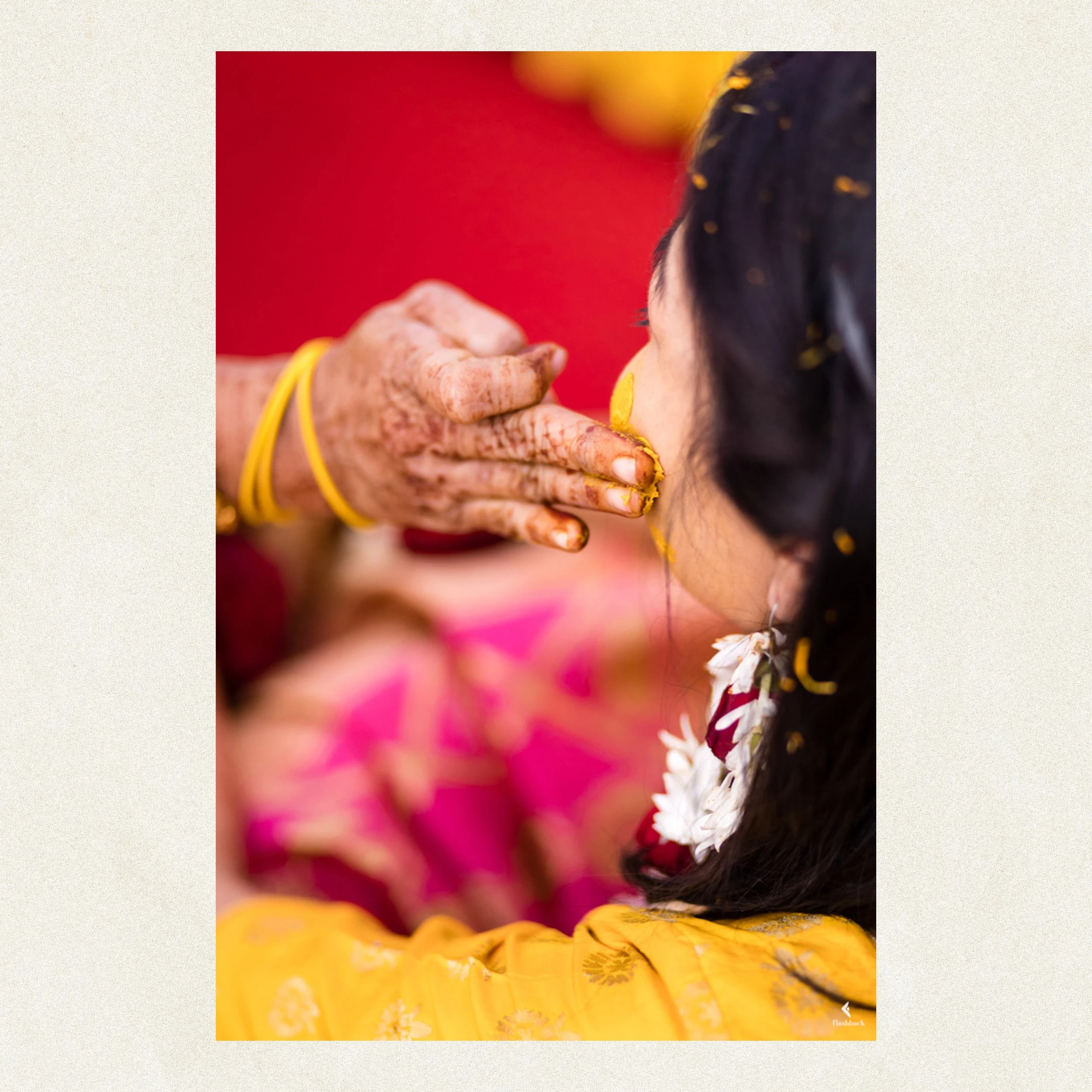 Elderly woman applying a yellow powder to a young woman’s face during a traditional celebration, dressed in colorful Indian attire with floral jewelry.