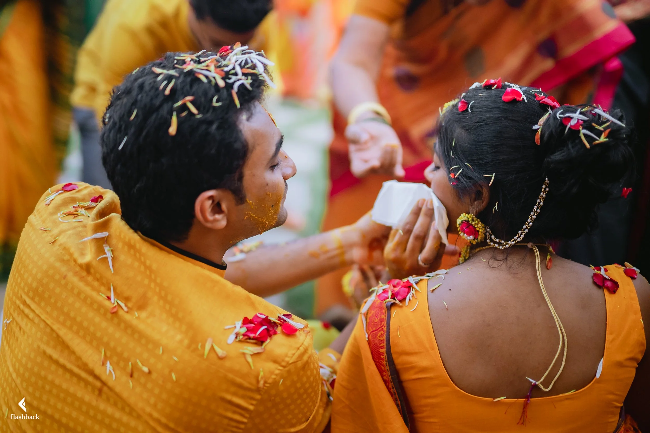 A traditional Indian wedding ceremony with a groom and bride dressed in yellow attire, participating in a ritual involving garlands and flower petals, surrounded by other people.