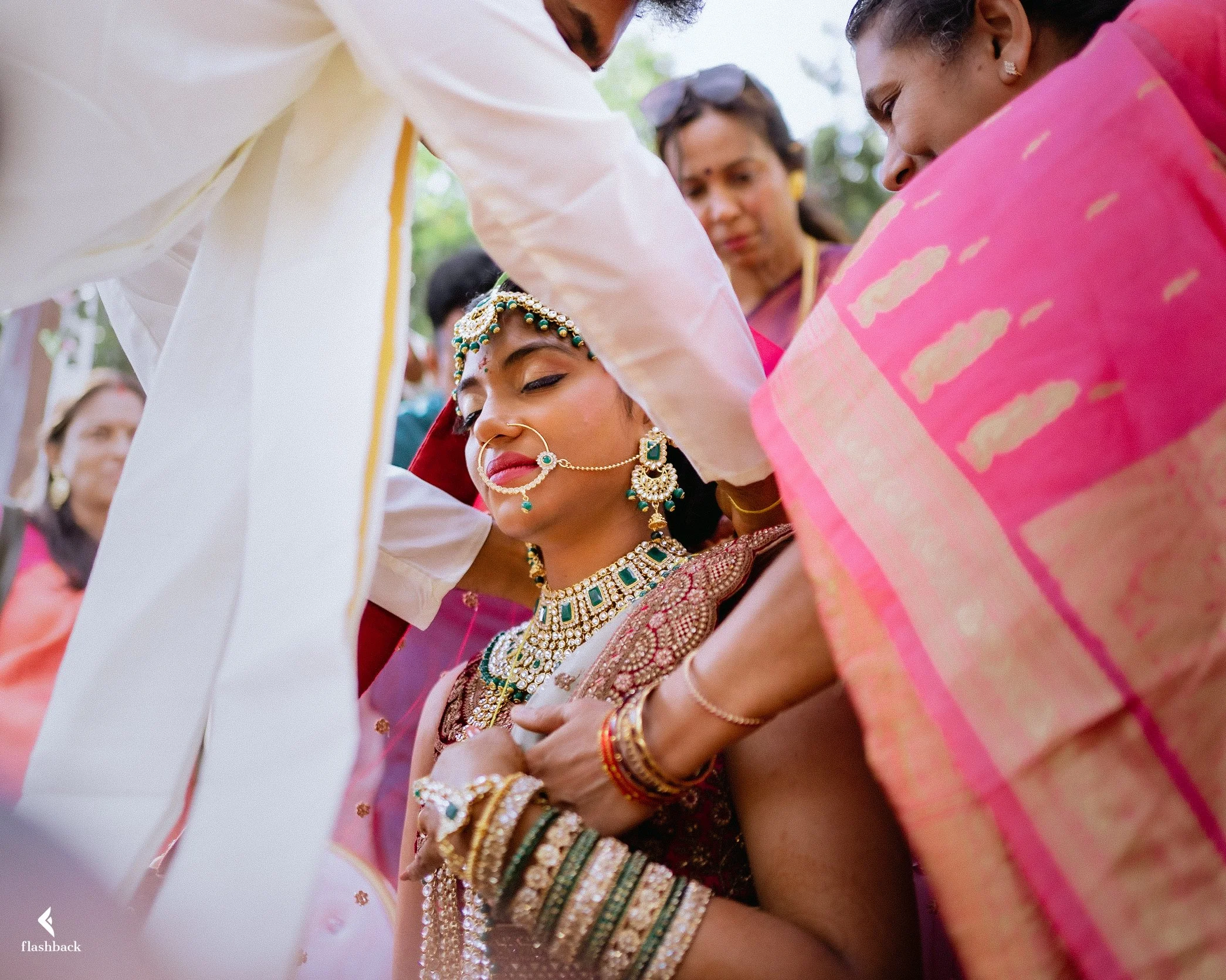 Indian bride wearing traditional jewelry and attire, surrounded by women at her wedding ceremony.