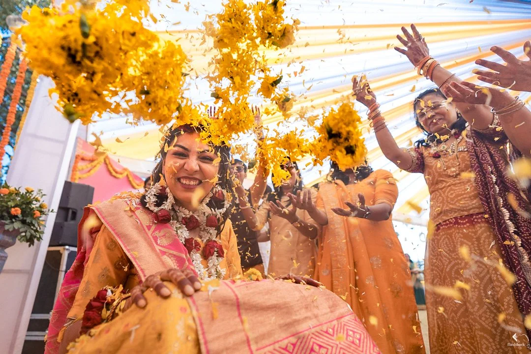 Celebrating a traditional Indian wedding, people dressed in colorful sarees throwing flower petals and smiling under a decorated canopy.