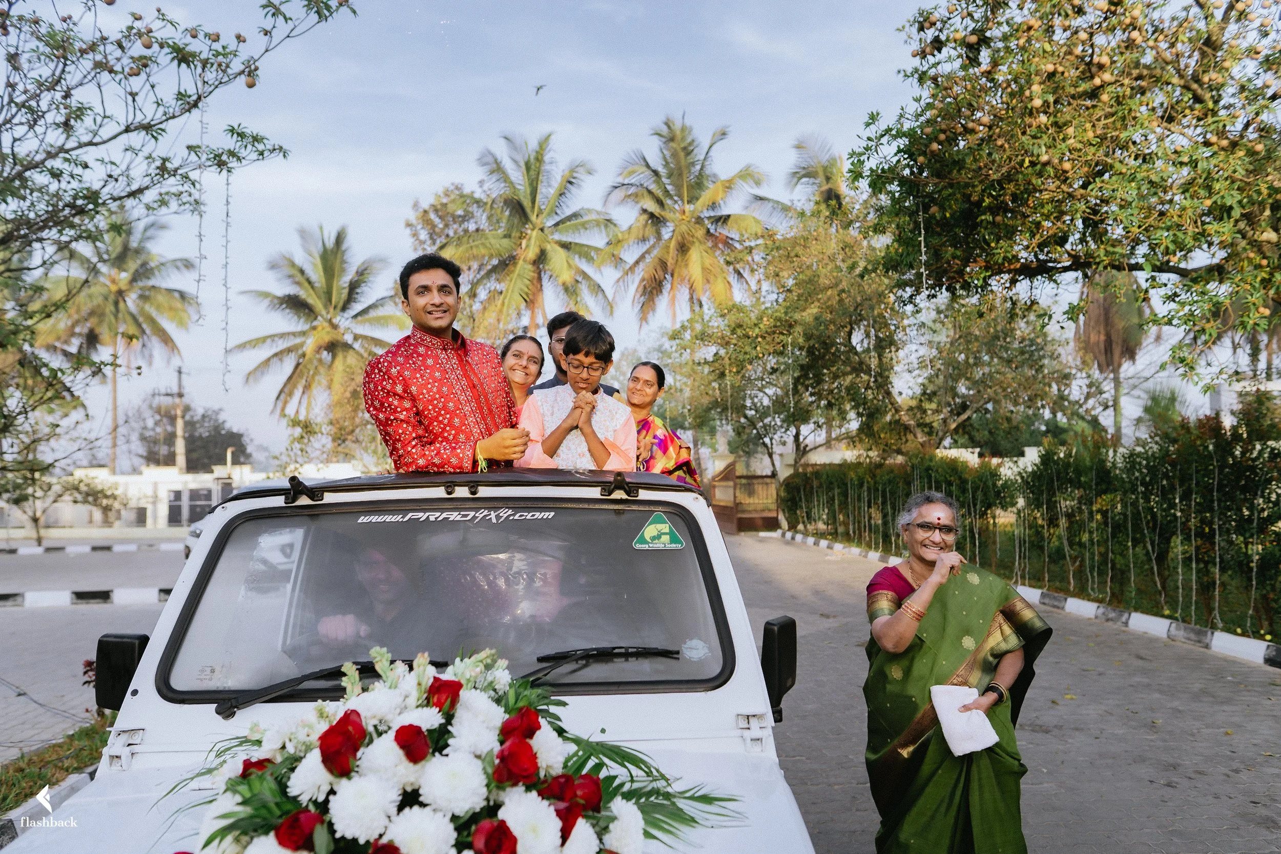 A group of people celebrating on top of a decorated white vehicle with flowers, near a woman in a green sari standing on the road, with trees and palm trees in the background.