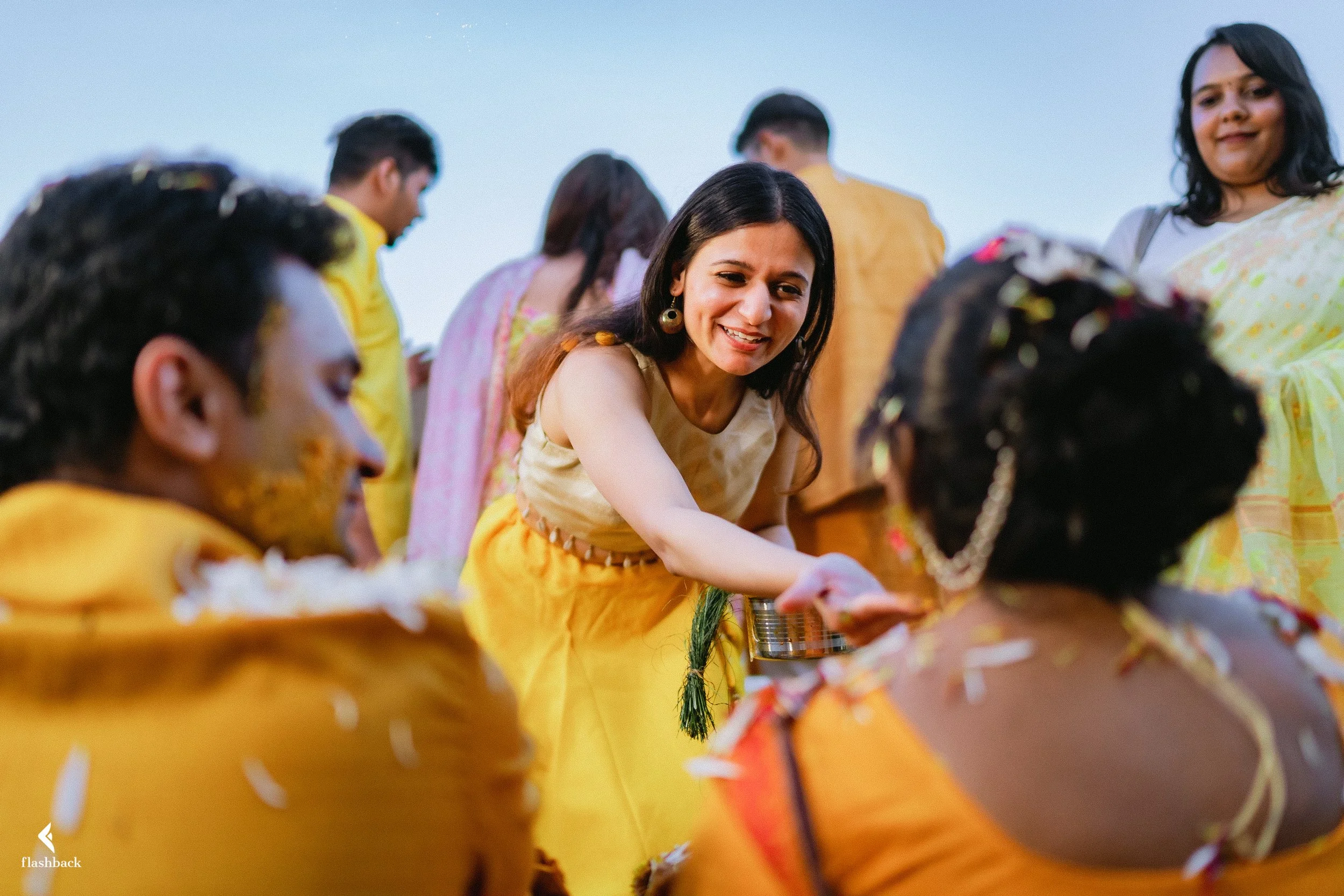 A woman in a yellow traditional dress is smiling and applying a cultural ritual to a woman with braided hair and flowers in her hair during a celebration outdoors, with other people in traditional attire in the background.