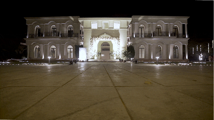 Illuminated large white mansion at night with arched windows and balconies, decorated with string lights and floral arches, with a wide paved courtyard in front.