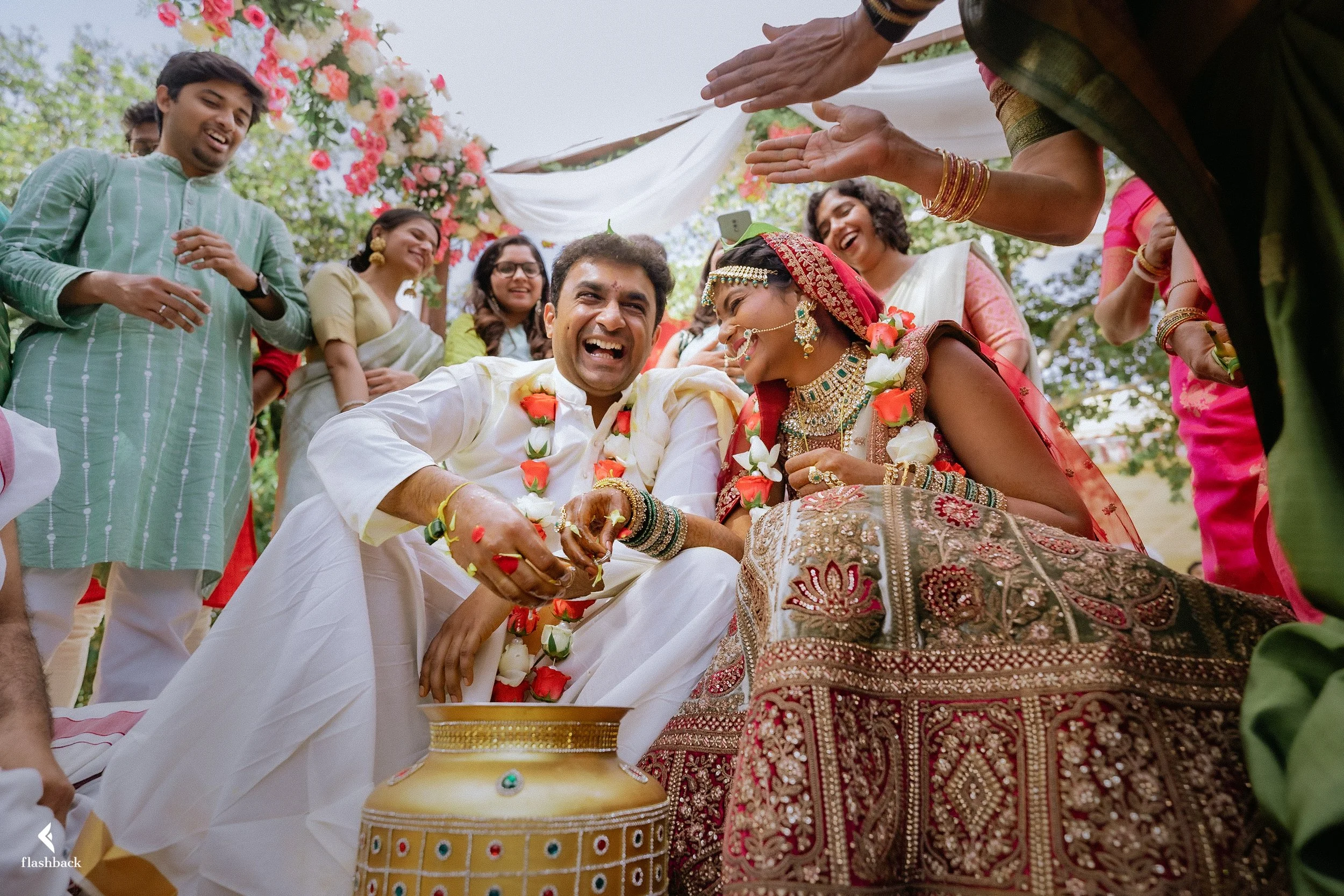A group of people celebrating an Indian wedding ceremony outdoors. The couple, dressed in traditional wedding attire, is surrounded by friends and family, with flowers and decorative drapes overhead. The groom is wearing a white kurta, and the bride