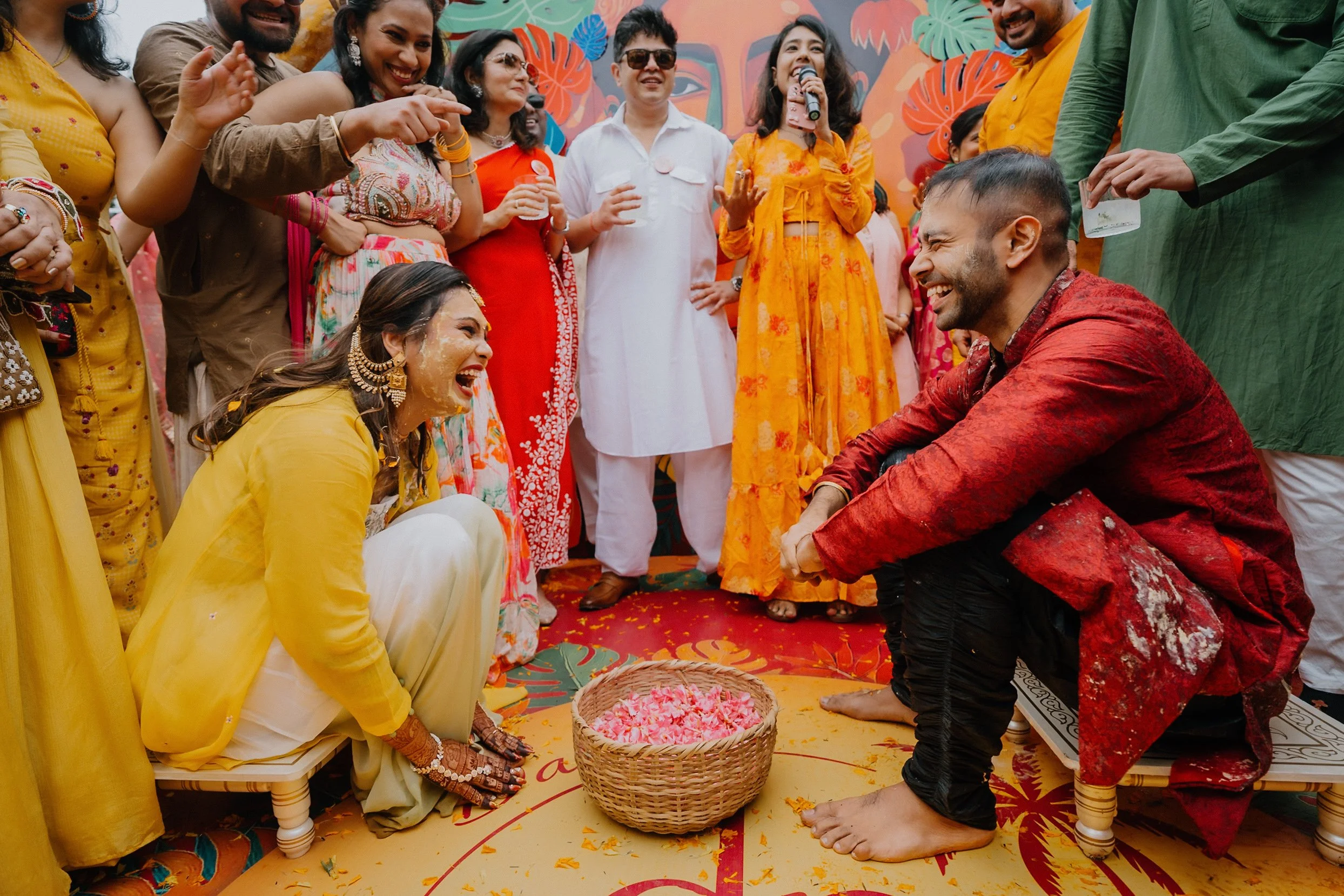 A joyful group of people celebrating an Indian wedding ceremony outdoors, with women in colorful sarees and men in traditional attire, participating in a ritual involving flower petals.