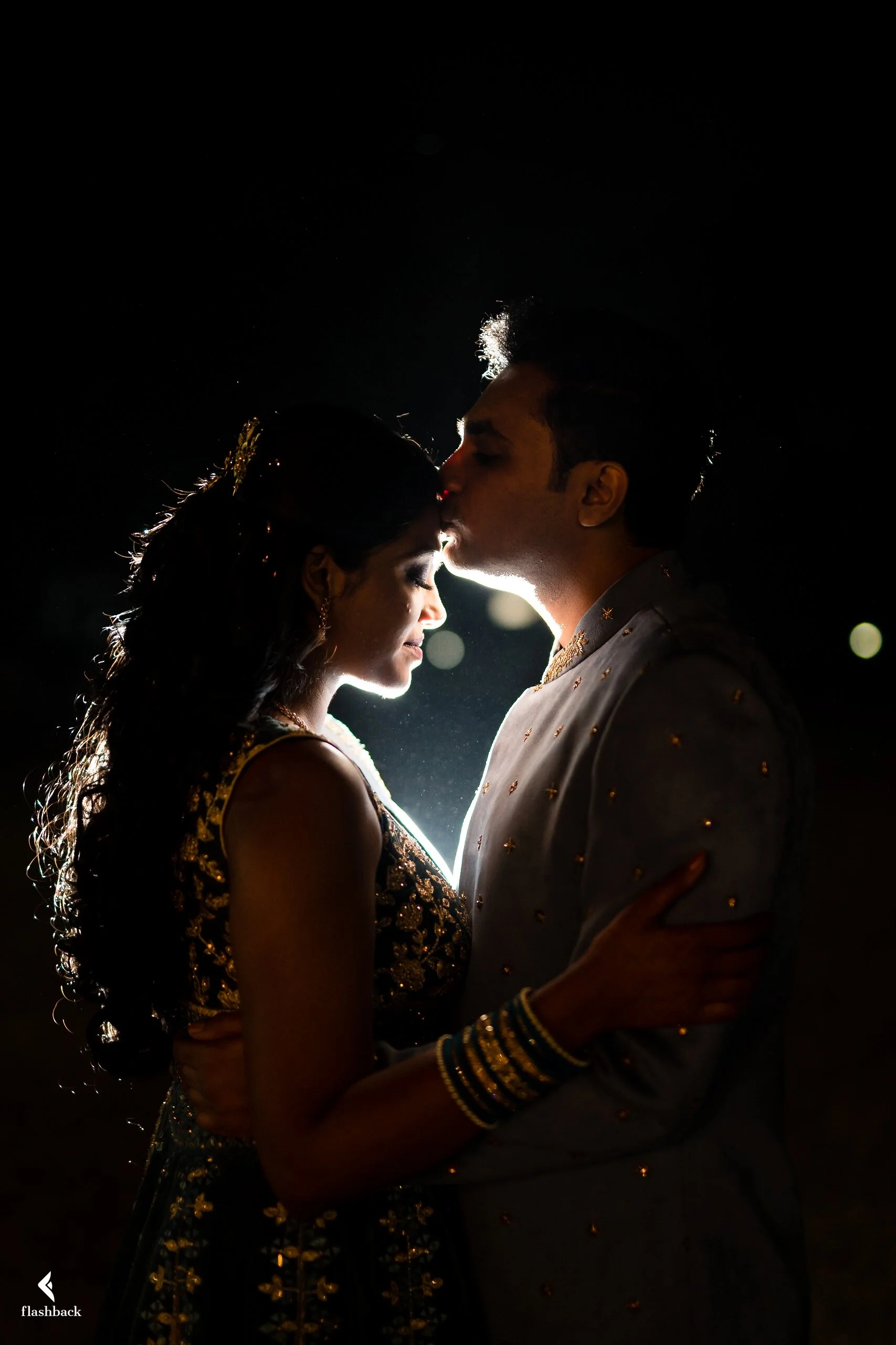 A couple is embracing and kissing at night, silhouetted against a dark background with backlighting highlighting their profiles, dressed in traditional Indian wedding attire.
