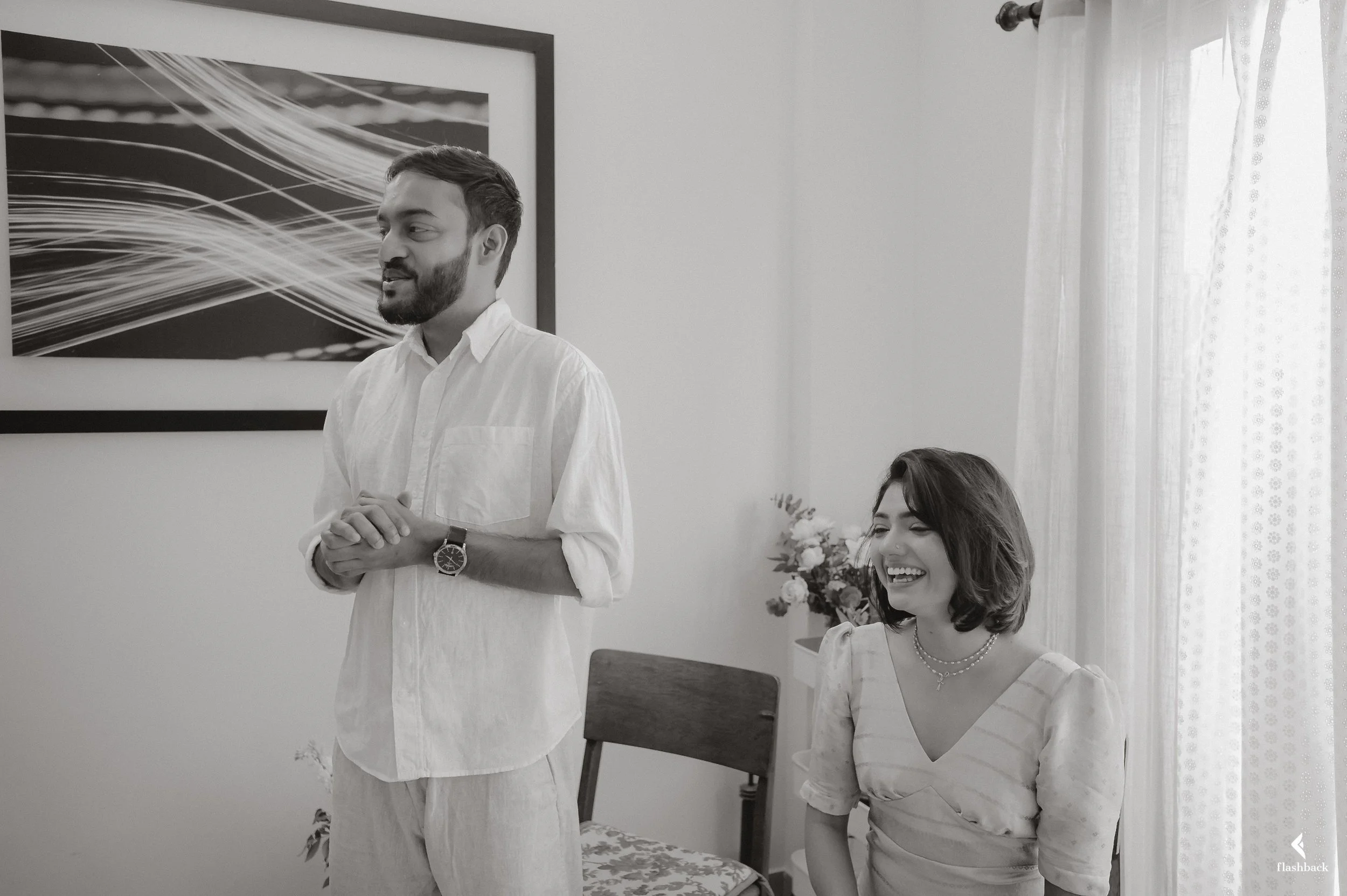 A man and a woman in a room, both smiling. The man is standing, wearing a white shirt and a watch. The woman is sitting, wearing a light-colored dress with striped pattern and jewelry. There is a framed artwork on the wall behind them and a chair wit