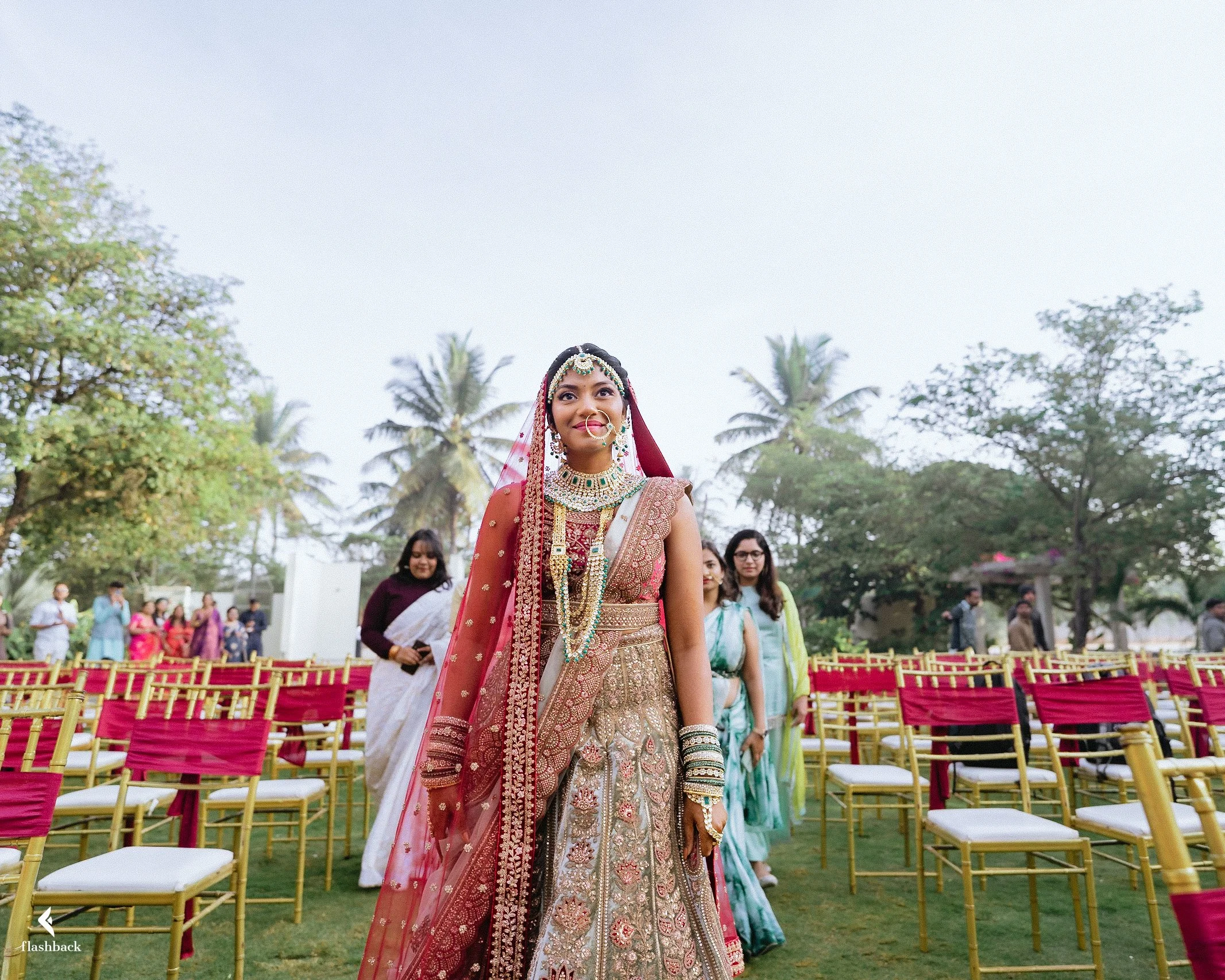 A bride in traditional Indian wedding attire standing outdoors among empty gold and pink chairs, with palm trees and guests in the background.