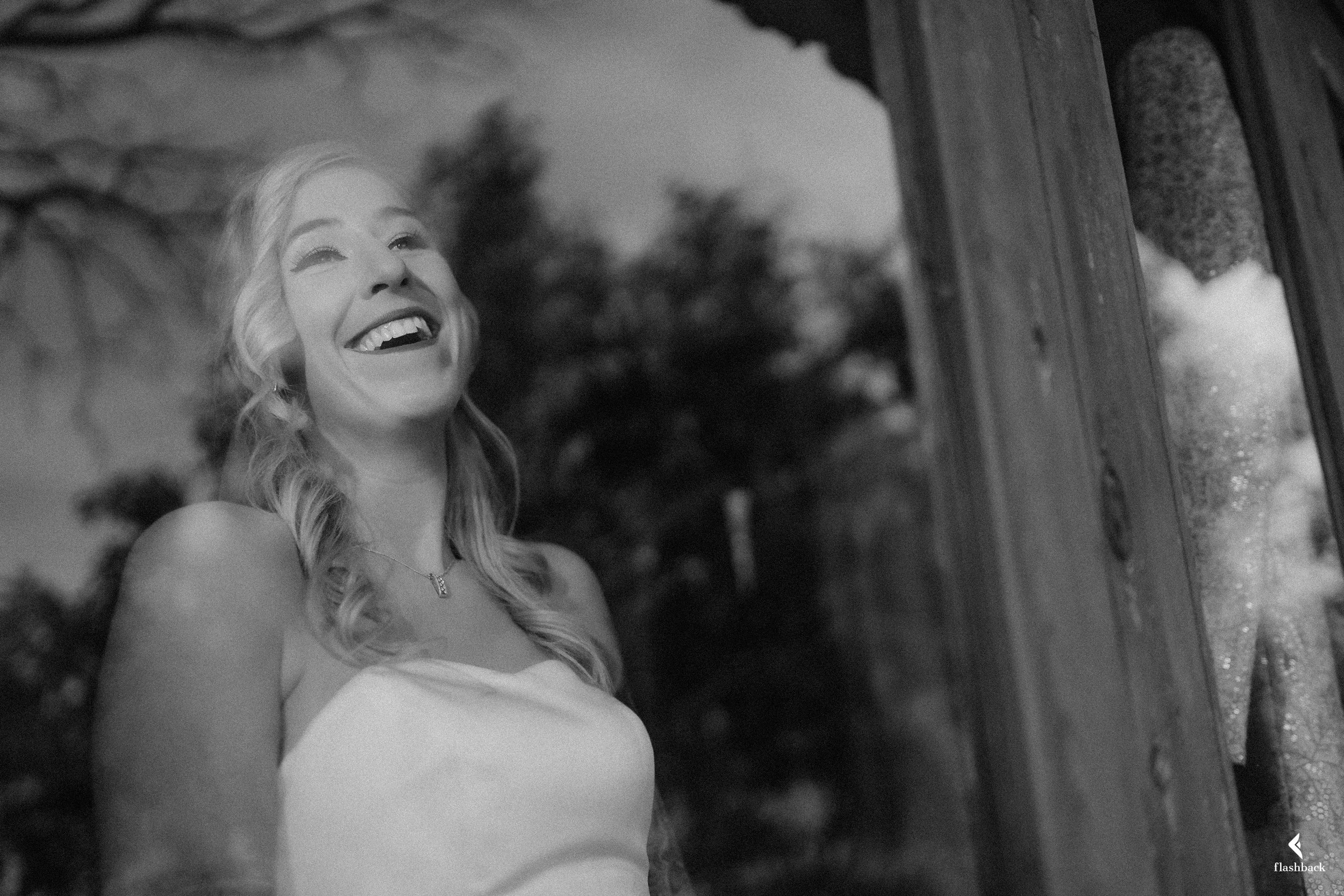 A smiling woman with wavy hair looking upward, reflected in a mirror, with trees and sky in the background.