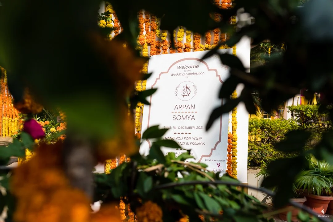 Wedding celebration sign for Arpan and Somya, dated December 6th and 7th, 2021, framed with orange and yellow marigold flower garlands and greenery, partially obscured by foliage.